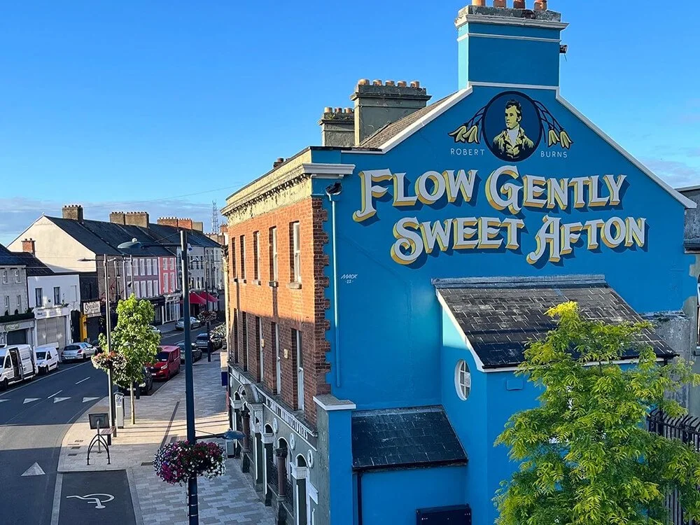 A street view with colorful buildings and a prominent blue building featuring the mural 'Flow Gently Sweet Afton' with a portrait of Robert Burns at the top.