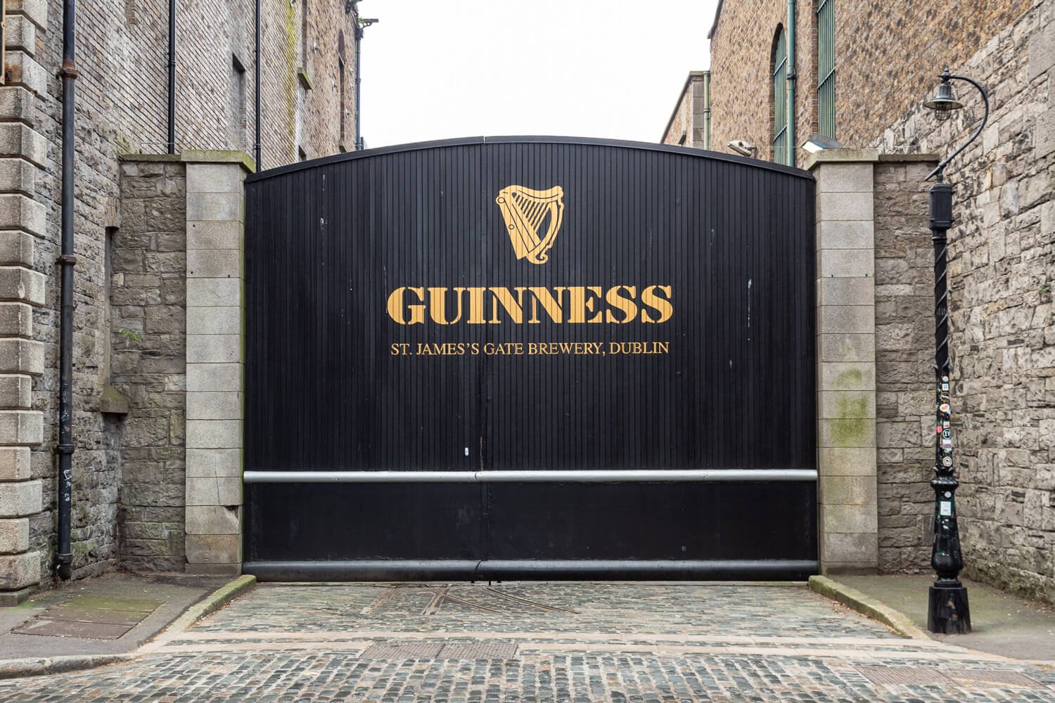 Black gate with gold Guiness logo and text, located between stone walls, marking St. James's Gate Brewery in Dublin.