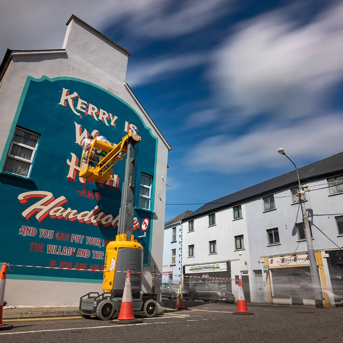 A cherry picker lifting workers to paint a mural on a building wall that says, "Kerry is W... and Handball." The building is white with a blue mural and is in an urban area with other buildings, parked cars, and a cloudy sky.