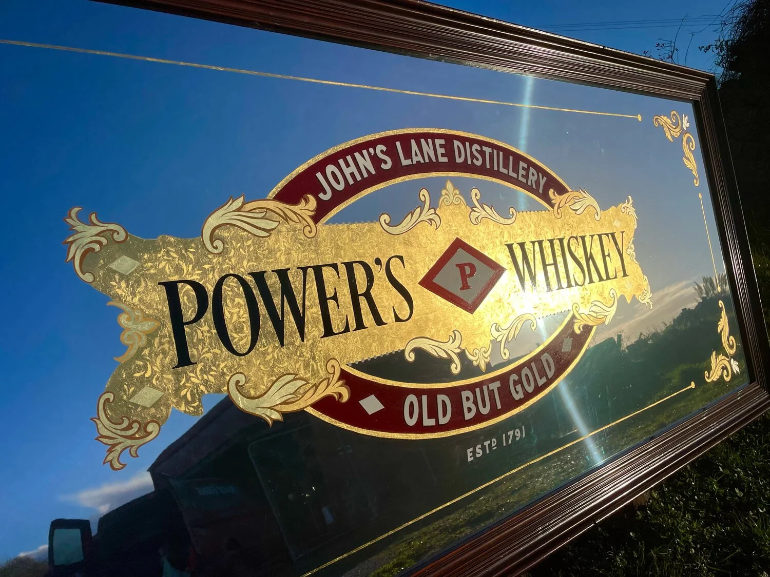 A decorative gold leaf sign for Powers Whiskey with gold and red accents, displaying the phrases 'Old but Gold' and 'Est 1791'.