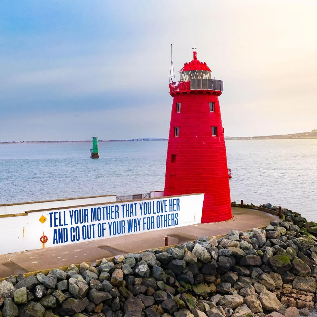 A red lighthouse situated on a rocky shoreline, with a white wall displaying a message that reads, 'Tell your mother that you love her and go out of your way for others.' In the background, a smaller green lighthouse is visible in the water, with calm ocean waters and a distant horizon.