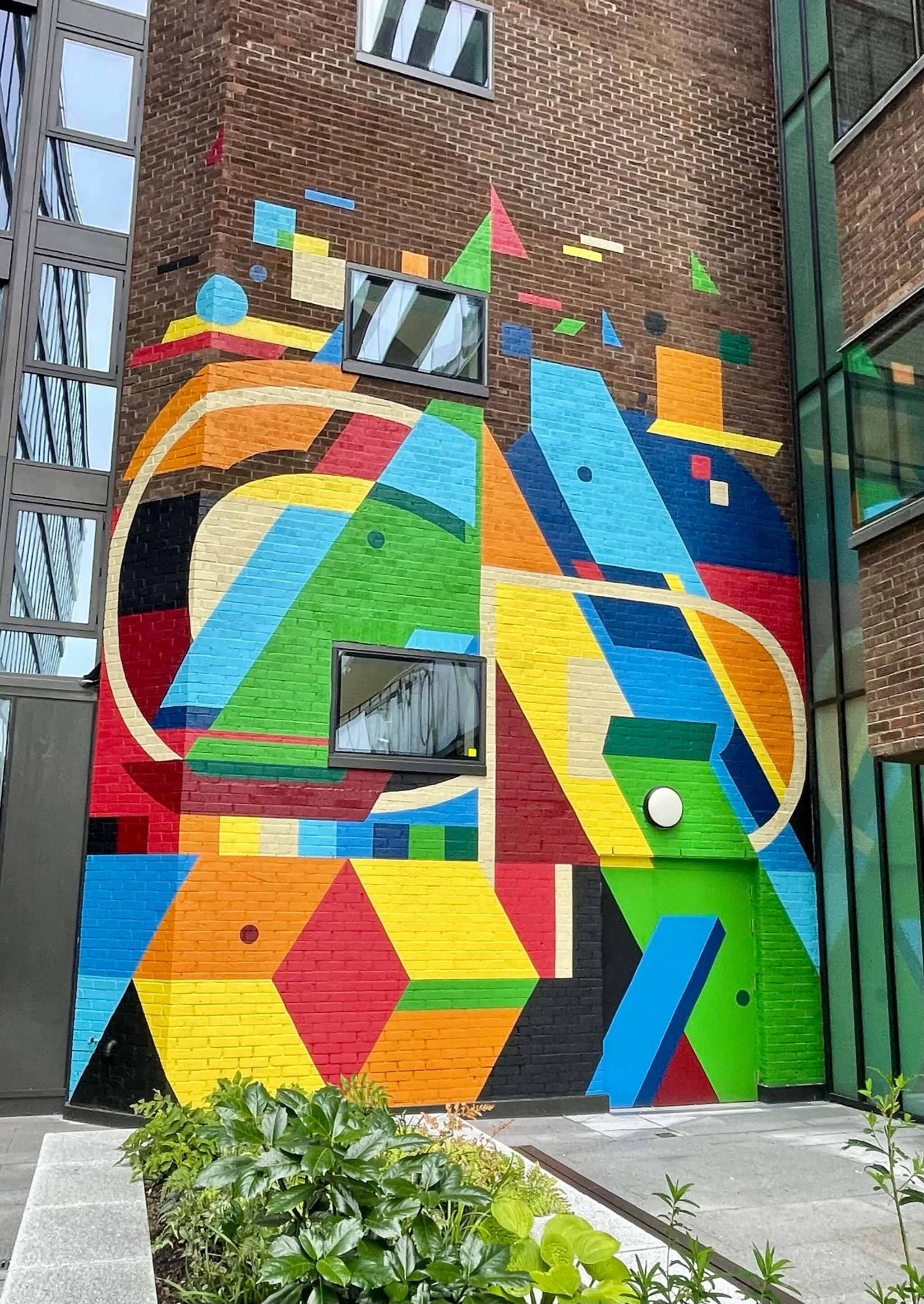 Colorful geometric mural on brick building with small plants in foreground.