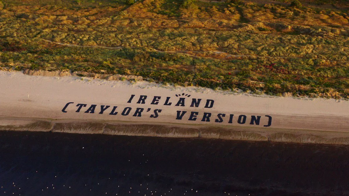 A beach with large text painted on the sand reading 'Ireland (Taylor's Version)' above the ocean with a grassy area beyond the beach.