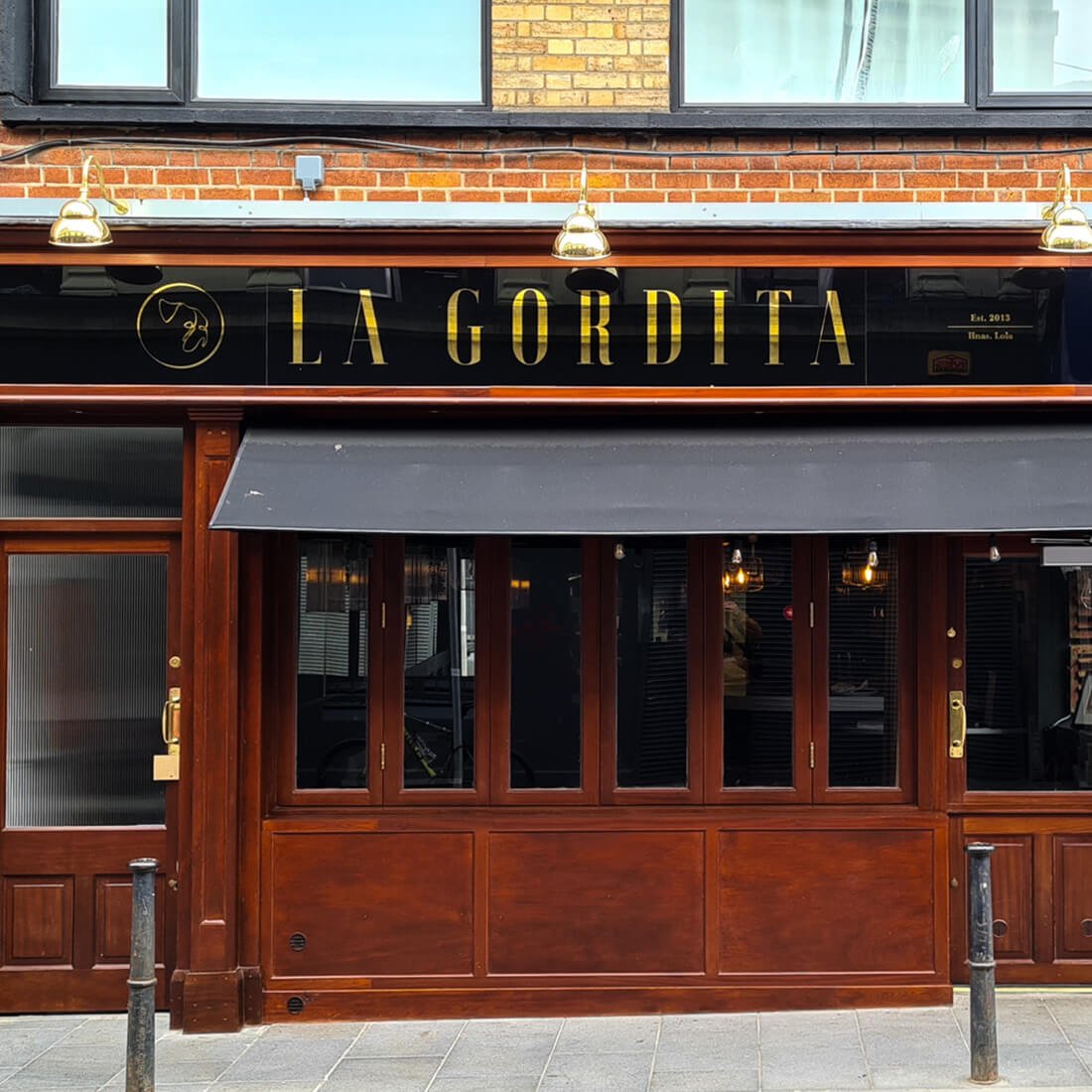 Restaurant storefront with black sign reading 'LA GORDITA' in gold letters, wooden door and windows, and brick and glass building exterior.