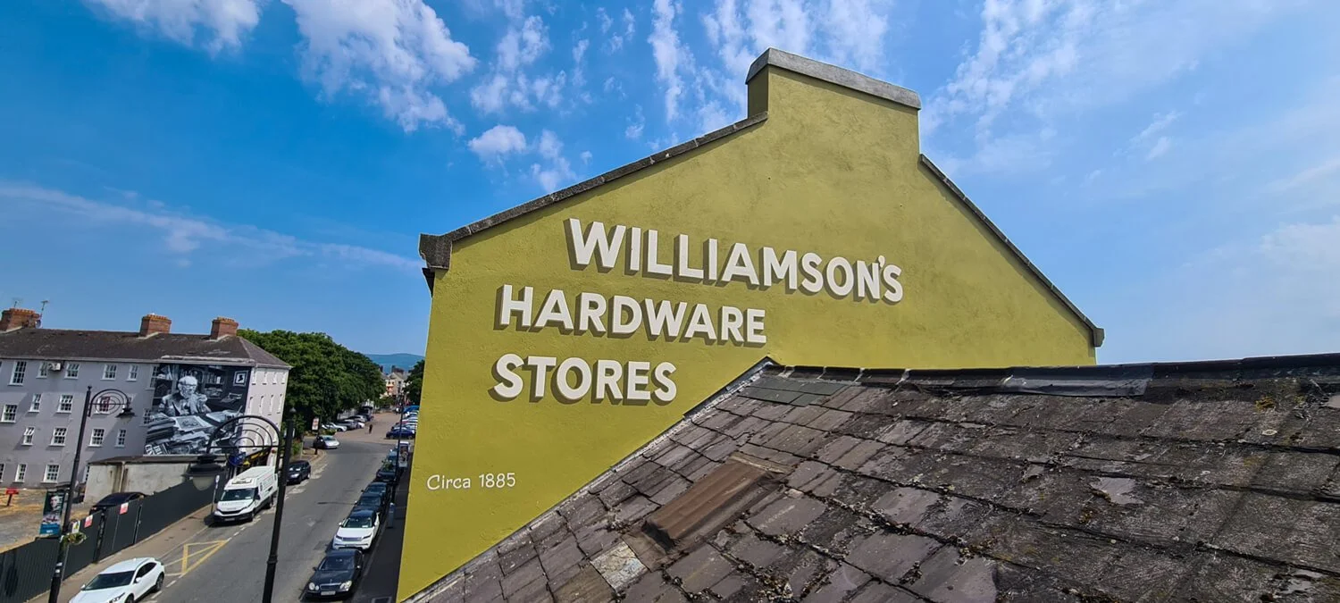 A large green building wall mural with white text that reads "Williamson's Hardware Stores" and "Circa 1885" underneath. The building has a steep roof with old, weathered shingles and is situated on a street with cars parked along the sidewalk.
