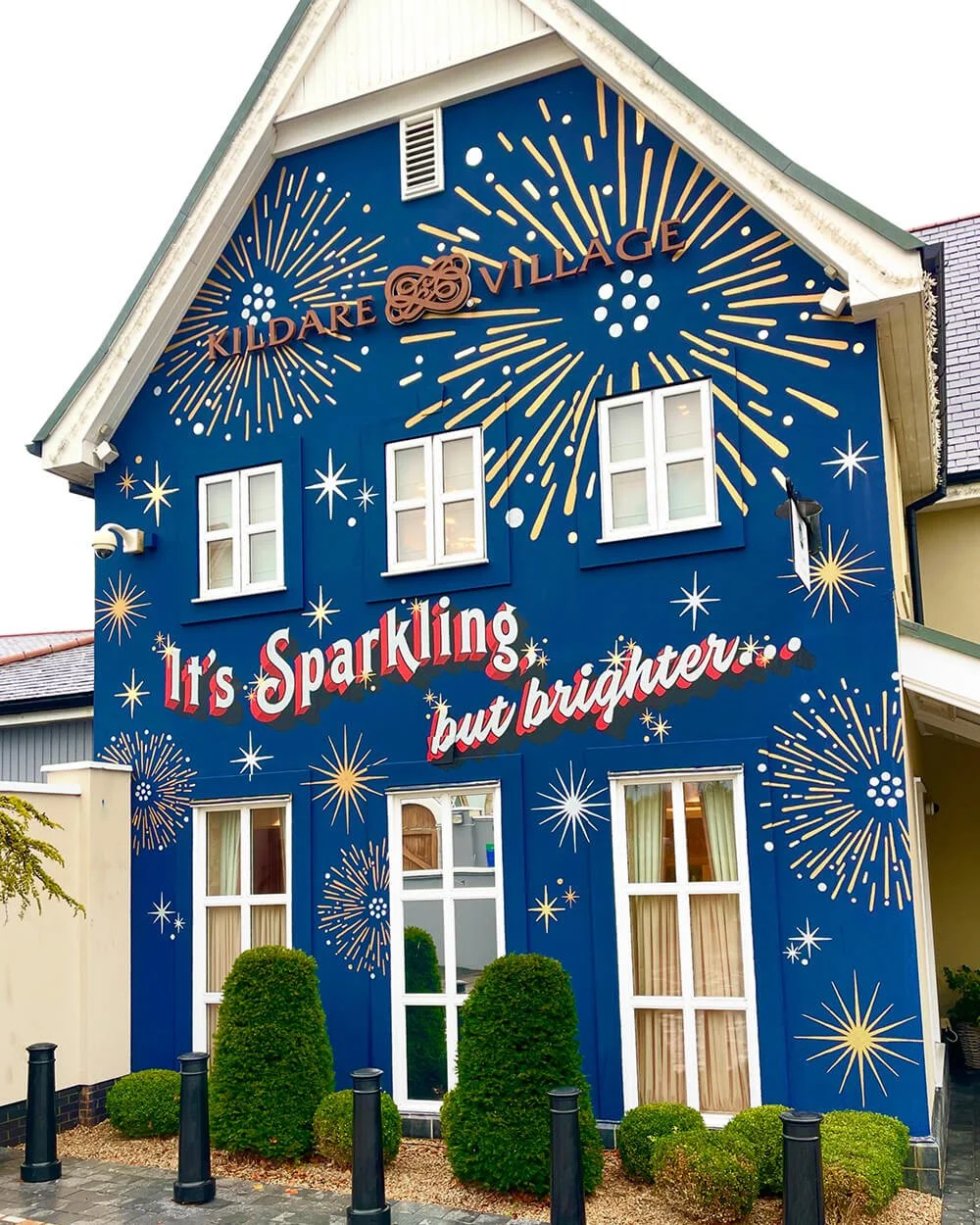 Colorful building painted dark blue with white and yellow fireworks and star designs, featuring a sign that reads 'Kilmore Village' at the top and a slogan 'It's Sparkling but brighter' in the middle. The building has several windows and a small land