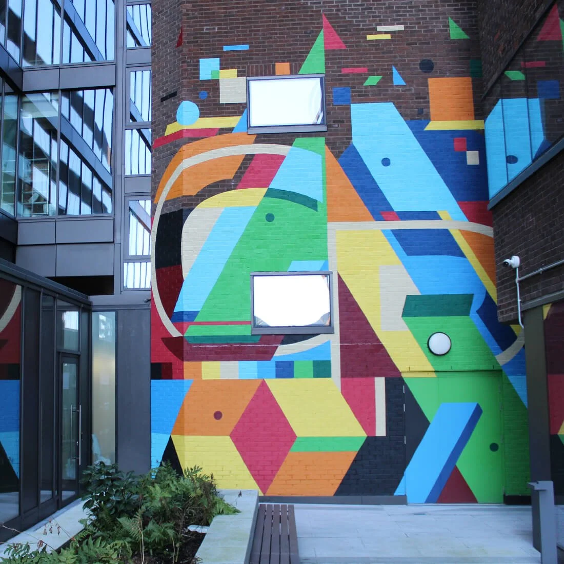 Colorful geometric mural painted on a brick building wall, featuring various shapes such as hexagons, squares, and triangles in bright colors including blue, green, yellow, orange, red, and black.