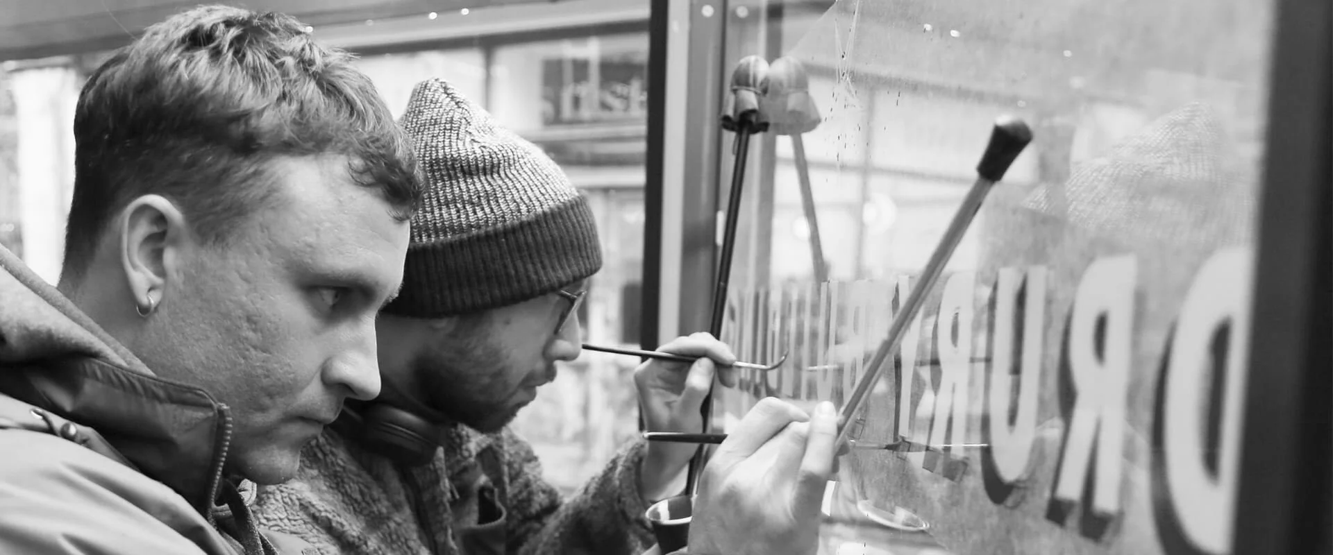 Two young men with short hair, one wearing a hoodie and the other a beanie, decorate a window with paint using brushes, with spray bottles and other tools nearby.
