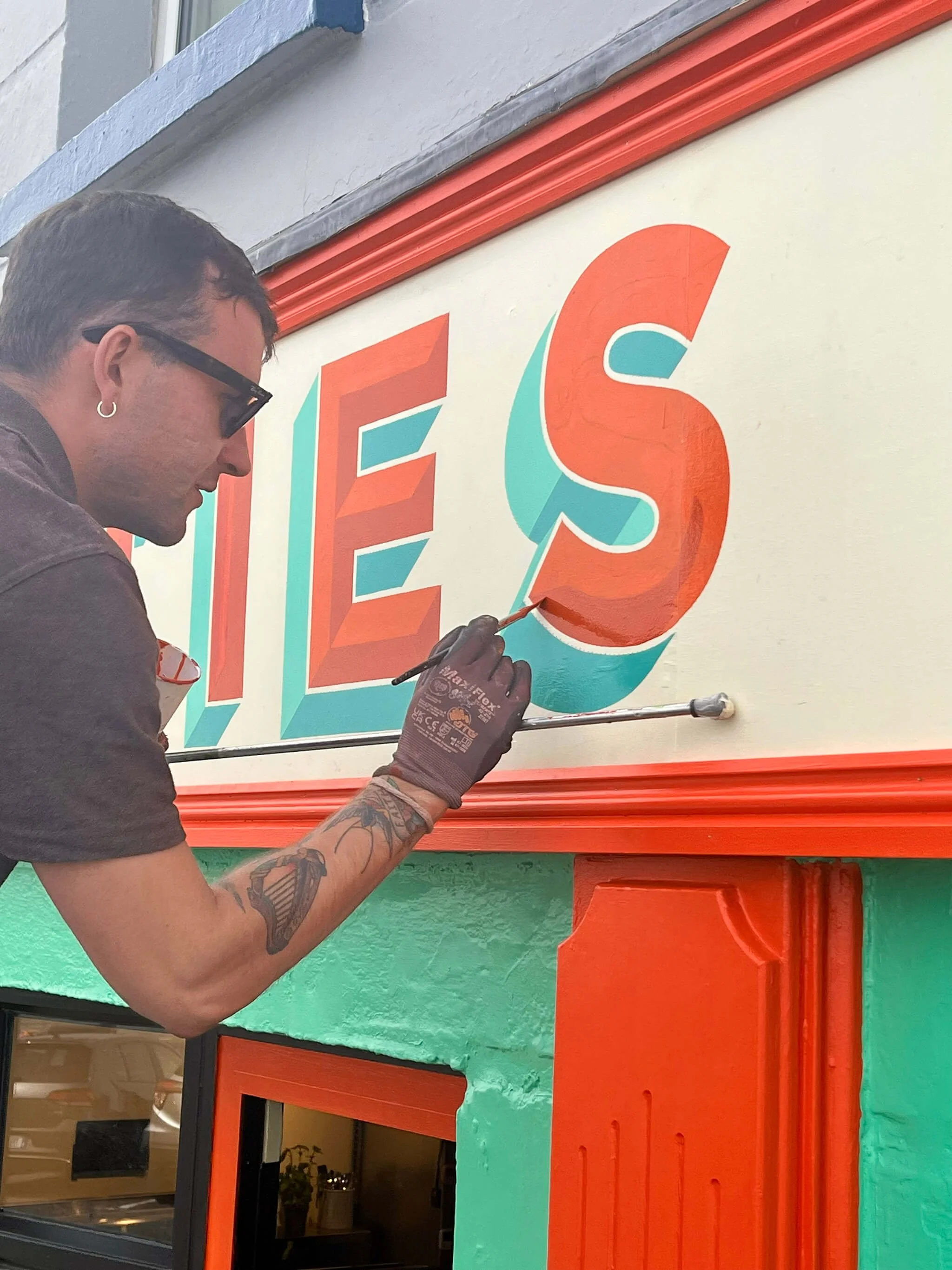 Cormac Dillon, man with tattoos, glasses, and a hoop earring is painting a colourful sign on the exterior wall of a building. He is using a small brush to add orange color to the letter 'S' in the sign.