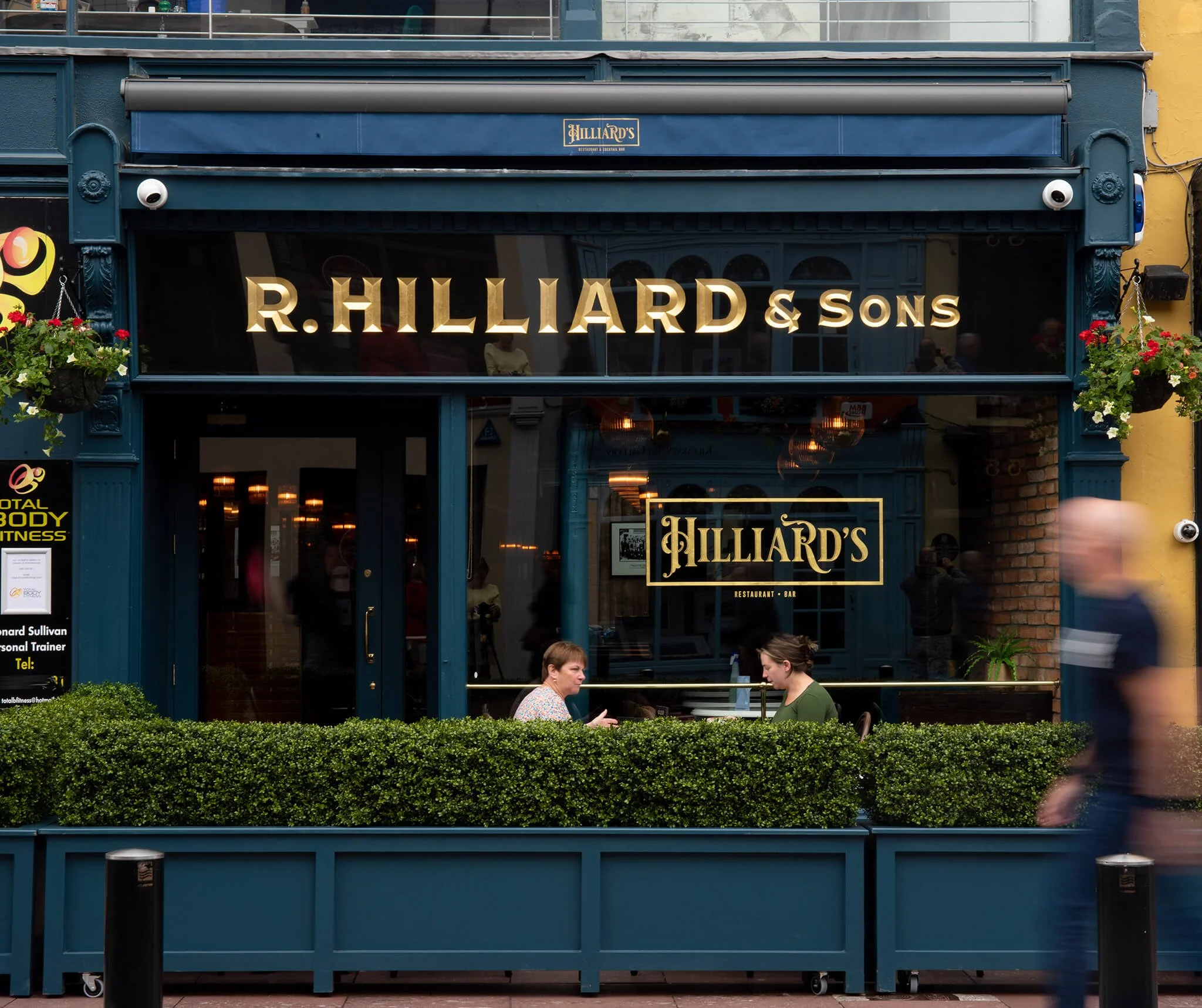 Exterior gold leaf signage of a restaurant named R. Hilliard & Sons with a glass window sign that reads Hilliard's Restaurant & Bar. Two women sit at a table outside, and a person is walking past.