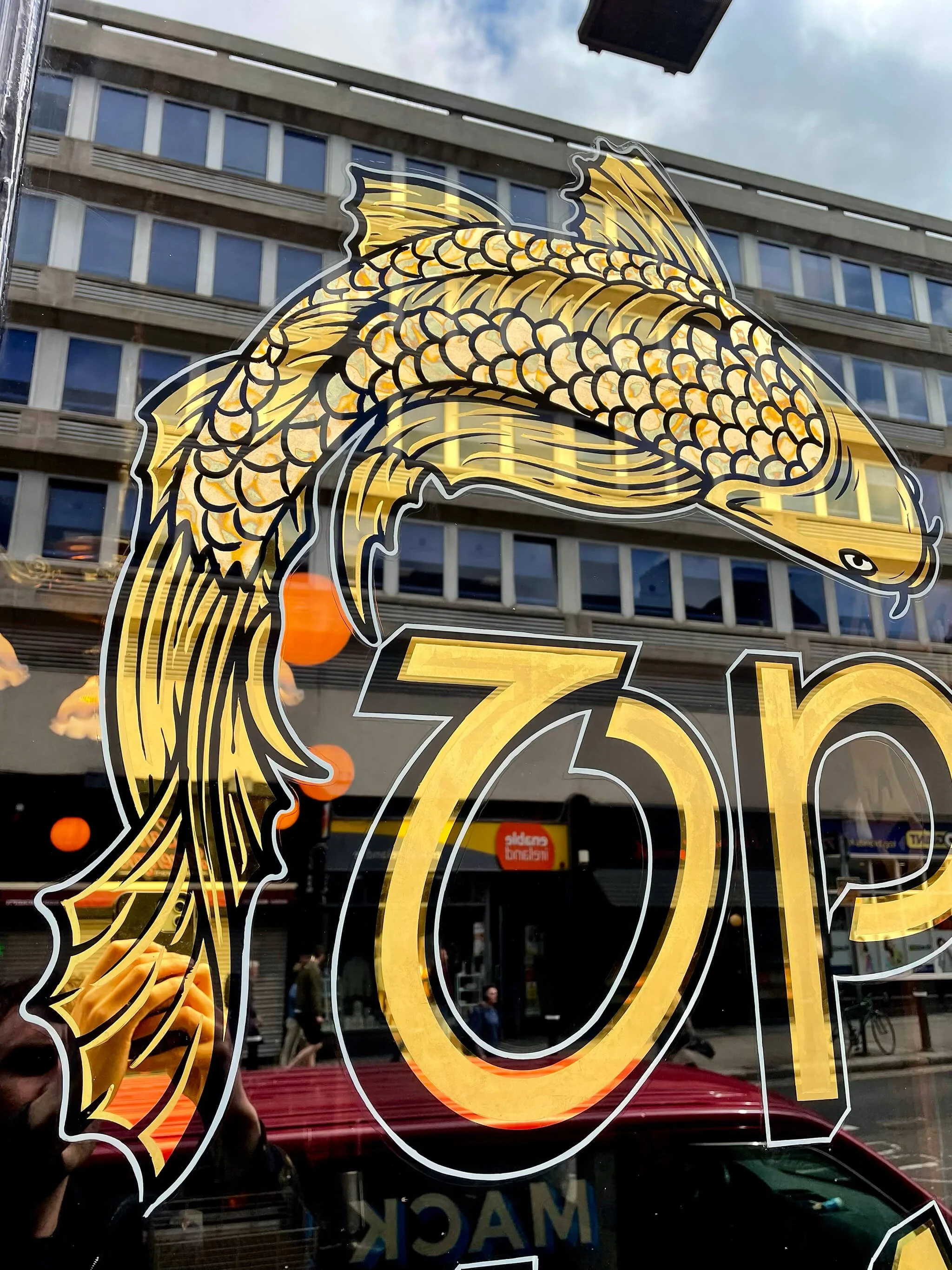 Design of a gold leaf koi fish in gold and black with the number 73N on a glass window, reflecting a city street scene with buildings and pedestrians.
