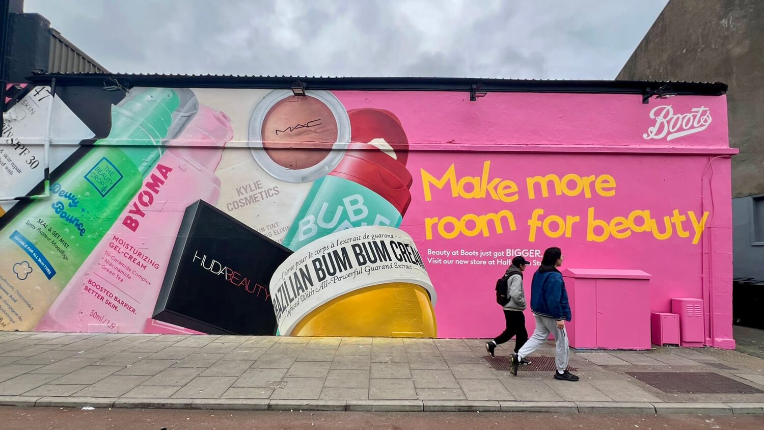 A large, colorful advertisement mural on a pink wall promotes beauty products at Boots, featuring various skincare and cosmetic bottles, with the slogan 'Make more room for beauty'. Two people walk past the mural on the sidewalk.