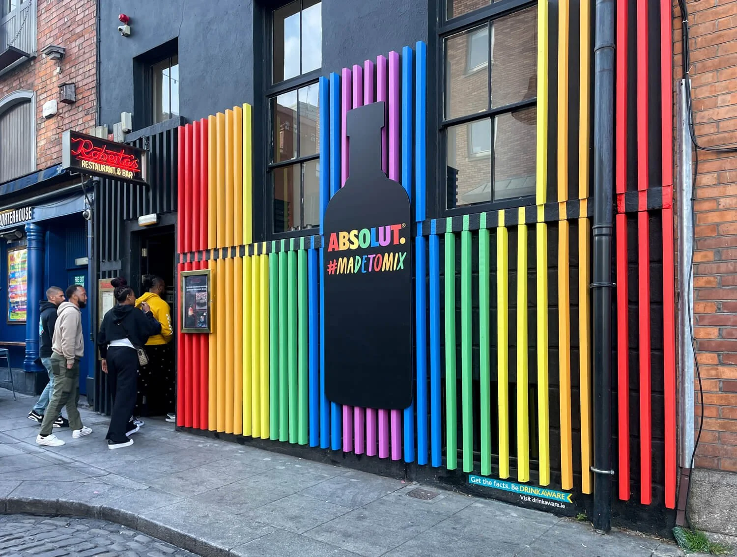 Colorful exterior of Roberta's restaurant in Dublin with rainbow vertical slats, a large bottle shape sign, and a group of people standing in line outside.