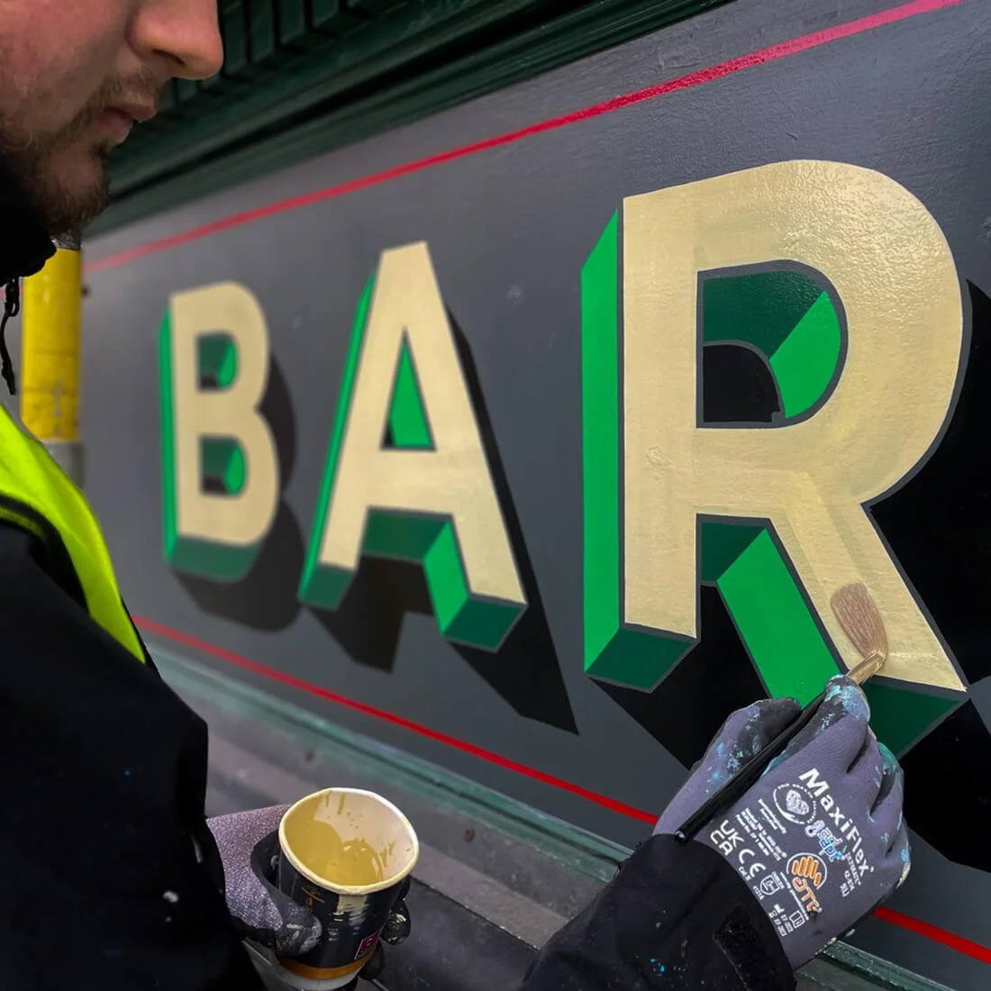 Person painting gold letters on a sign that spells out 'BAR' with a small paintbrush, wearing a high-visibility vest and gloves.