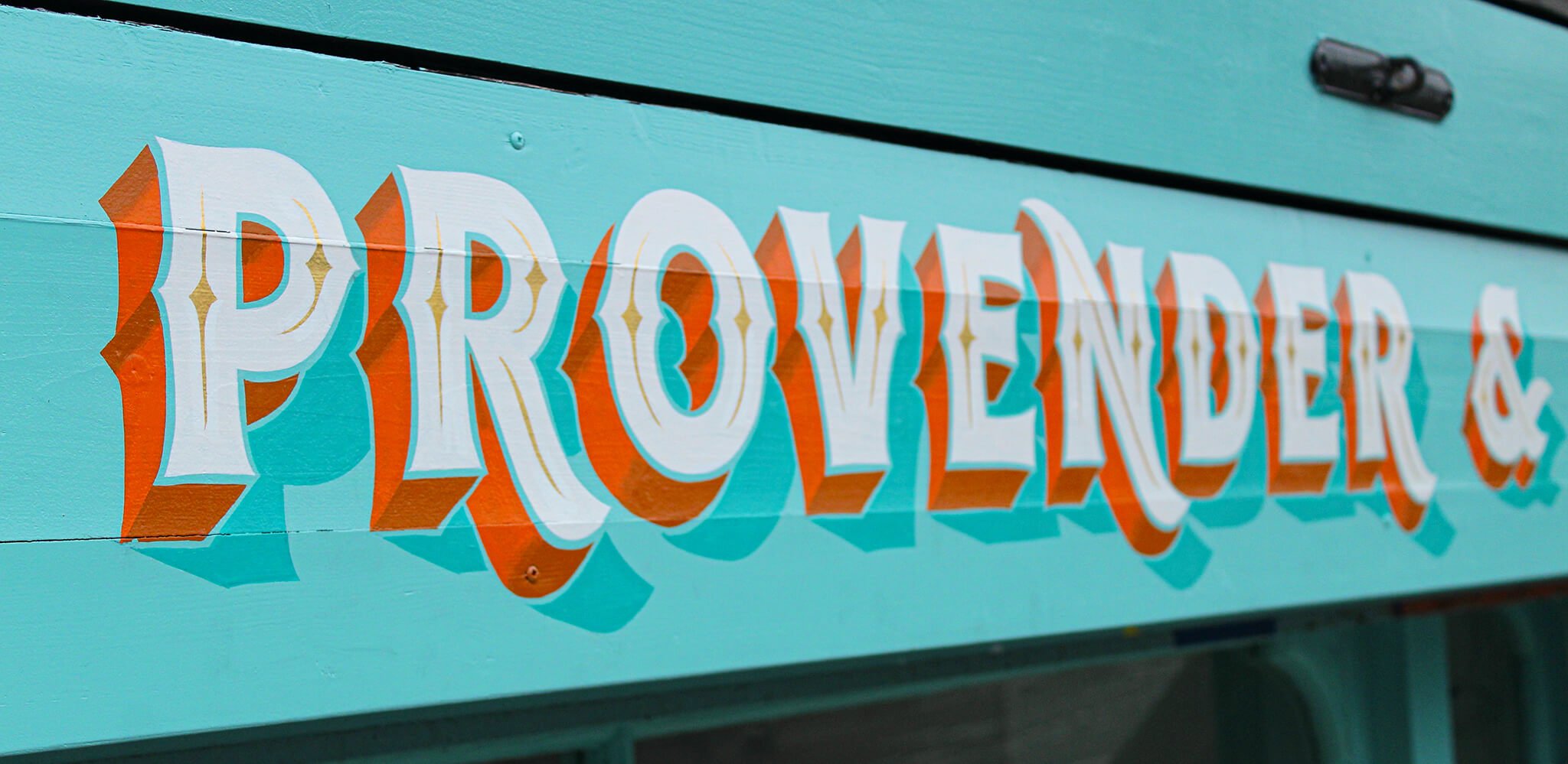 Colorful sign with the words 'PROVENDERS' painted in bold white letters with orange shadowing on a turquoise wooden background.