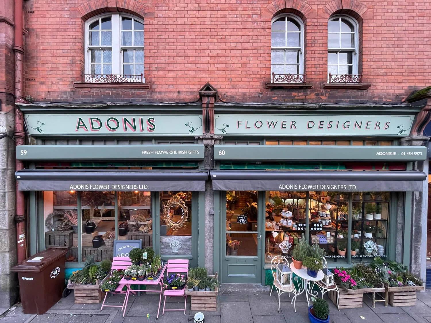 Exterior of a flower shop called Adonis Flower Designers, located on a brick building with two large windows above. The shop displays potted plants and flowers outside, with pink chairs, a small table, and decorative items. Signage indicates it sells