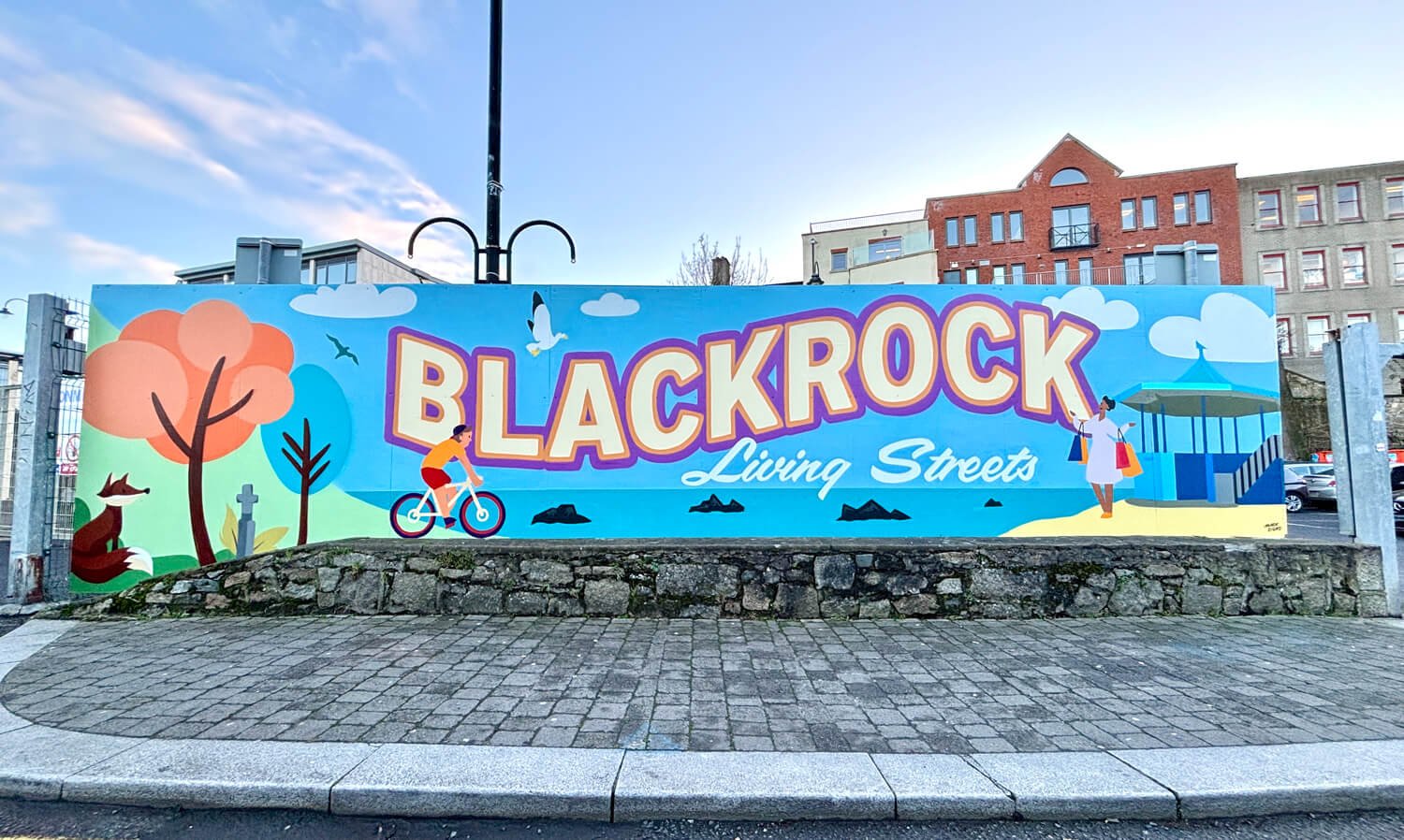 Colorful city mural advertising Blackrock Living Streets with illustrations of a cyclist, a woman walking with shopping bags, trees, birds, and a beach area.