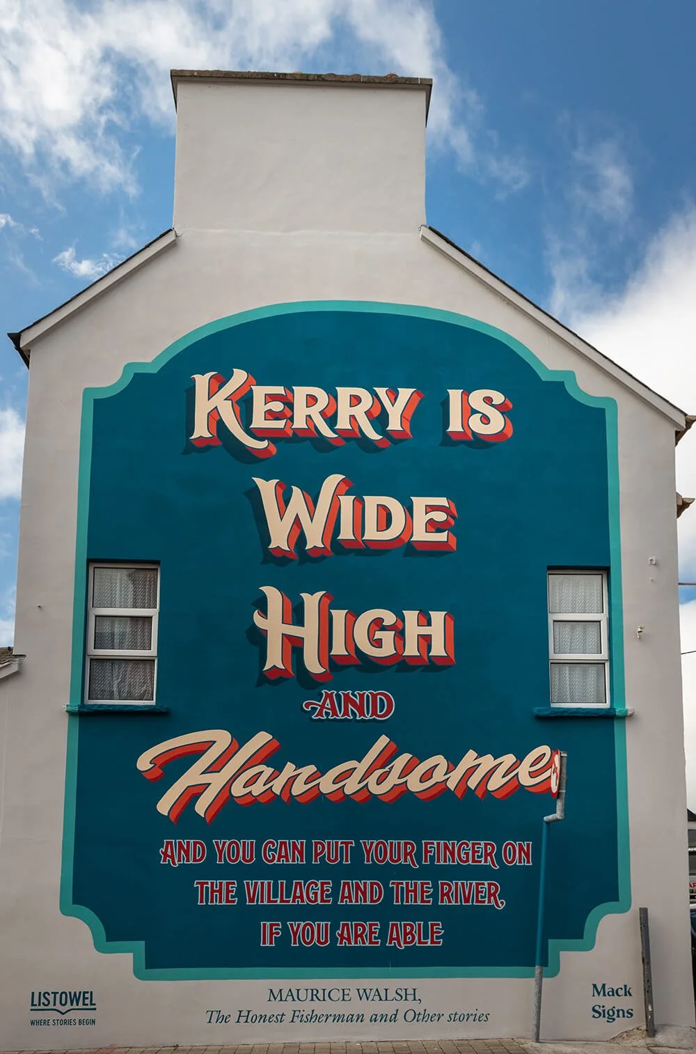 A large exterior wall with a colorful sign that reads "Kerry is wide high and handmade and you can put your finger on the village and the river if you are able." The sign is part of a pub called Listowel, with a quote from Maurice Walsh's story "The 