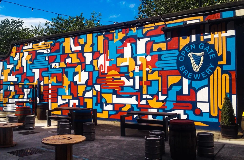 Colorful mural on an outdoor wall of Open Gate Brewery, featuring geometric shapes in red, blue, yellow, and white. Outdoor seating includes wooden tables and barrel-shaped stools.