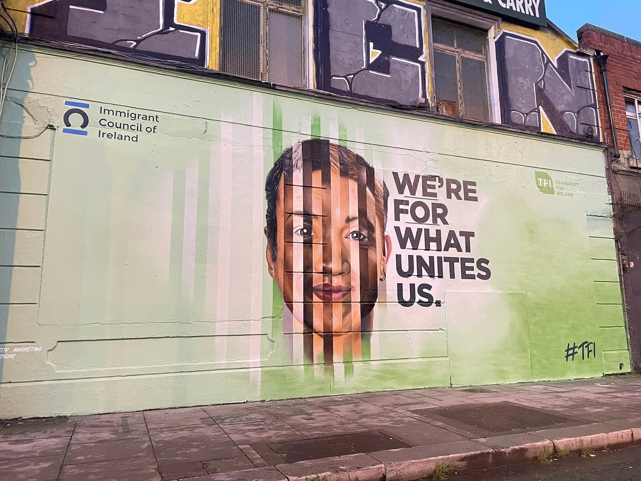 Mural of a woman's face with the text 'WE'RE FOR WHAT UNITES US' and logos of the Immigrant Council of Ireland and Transport for Ireland on a light green background.
