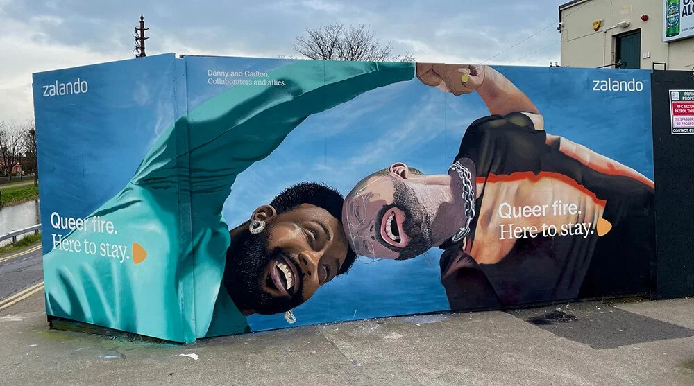Mural painting on a wall featuring two people, one with dark skin and short hair, and the other with light skin, a beard, and sunglasses. They are smiling and holding hands above their heads, with the words "Queer fire. Here to stay." written on it. 