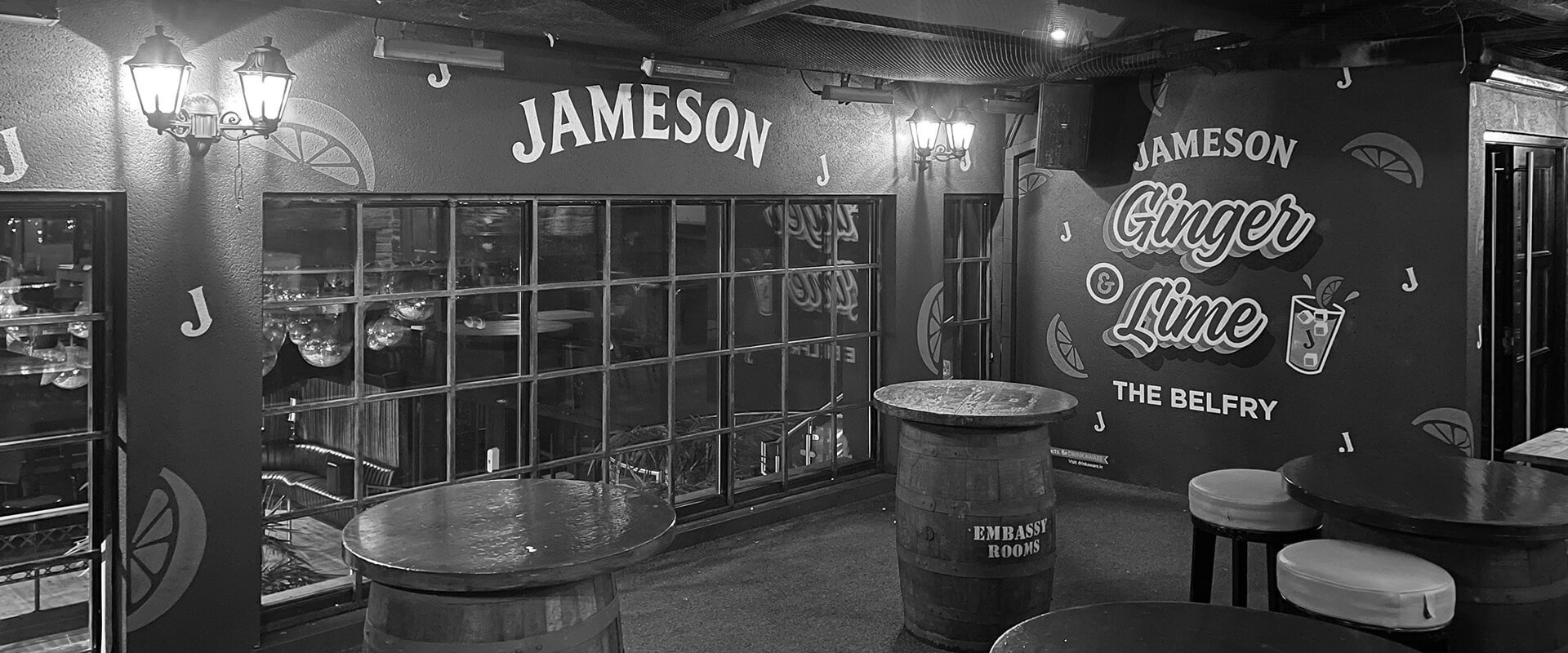 Inside a bar or pub with Jameson branding, tables, stools, and decorative wall with text indicating Ginger & Lime drink and the Belfry. There are windows and wall-mounted lights.