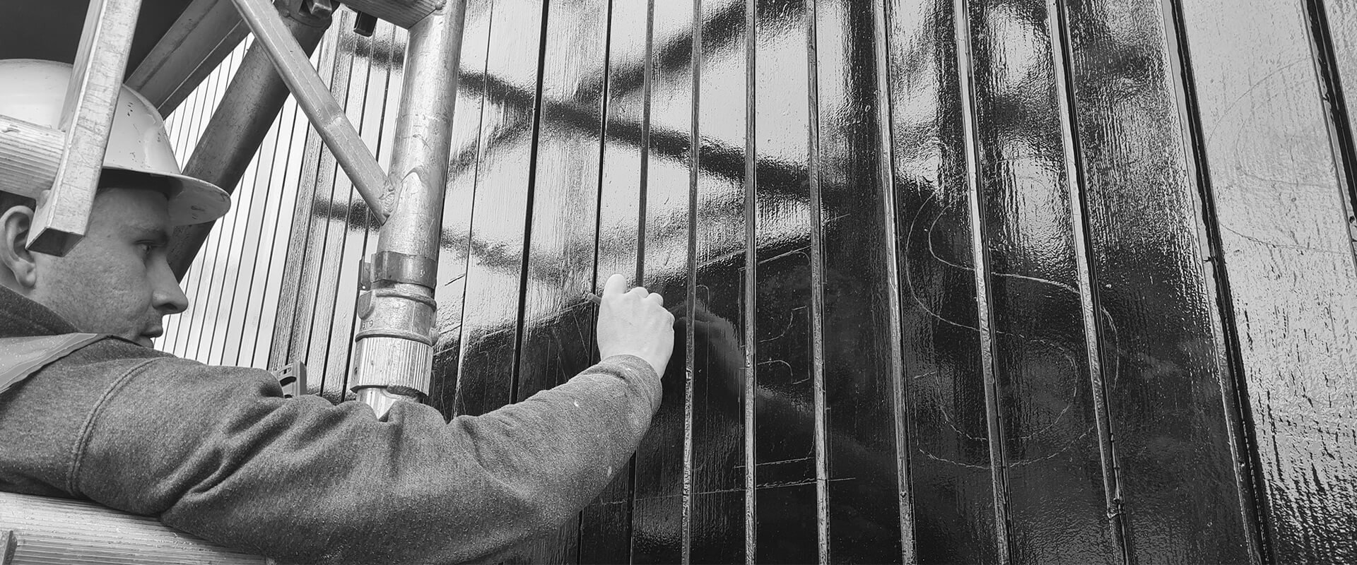 A worker with a hard hat and gloves is painting vertical metal bars black from a scaffold.