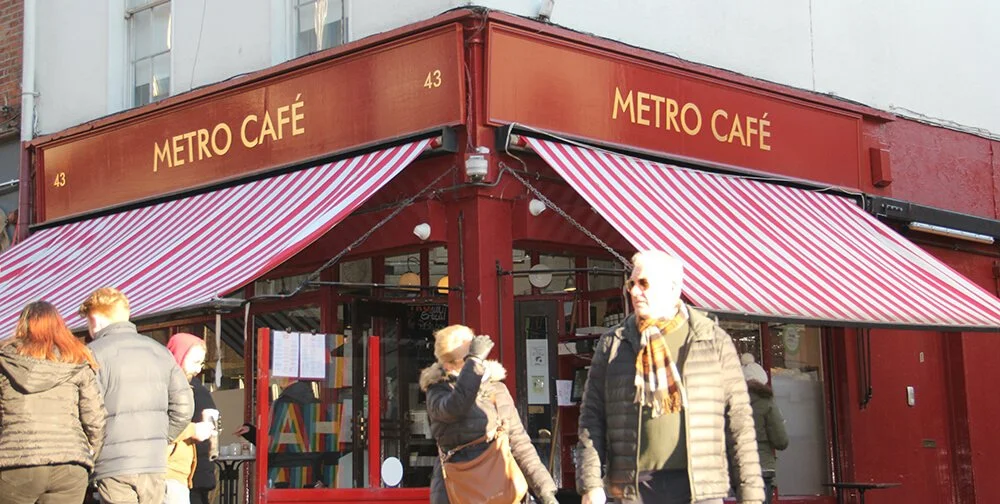 People walking past and entering Metro Café, a red-brick café with striped red and white awnings and gold lettering on a city street.