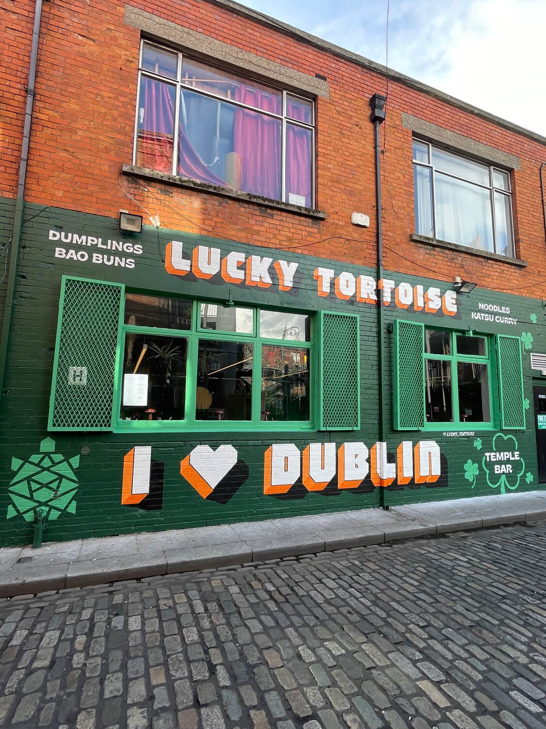 Exterior of Lucky Tortoise restaurant with green window shutters, colorful signage that says 'Lucky Tortoise' and 'I ♥ Dublin,' featuring shamrocks and food type references like dumplings, bao buns, noodles, and katsu curry.
