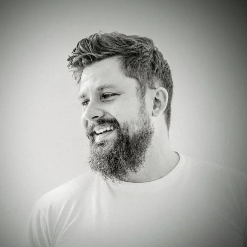 A smiling man with a beard and short, wavy hair, looking off to his left, wearing a plain white shirt, in black and white.