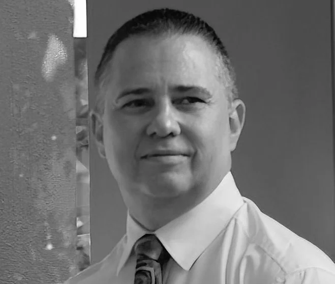 A black and white portrait of a man wearing a collared shirt and tie, smiling slightly, with a blurred background.