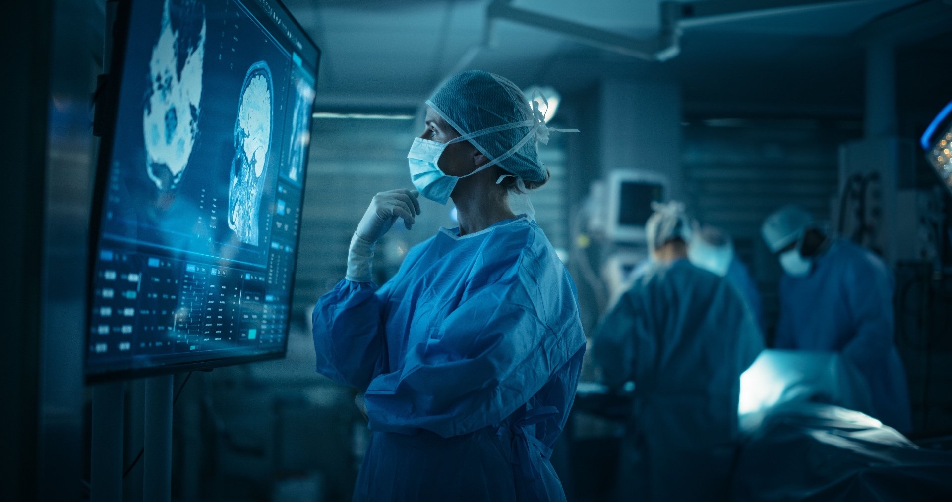 A medical professional in surgical attire, including a face mask and hair covering, looks thoughtfully at a large monitor displaying medical imaging in an operating room with other staff in the background.