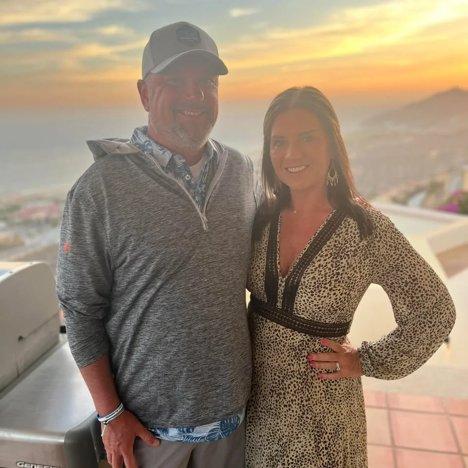 A man and woman standing together outdoors during sunset, with a scenic view of the ocean and coastline in the background.