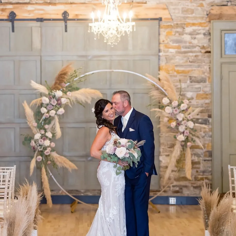 A bride and groom share a kiss during their wedding ceremony in a decorated indoor venue with a floral arch, chandelier, and wooden accents.
