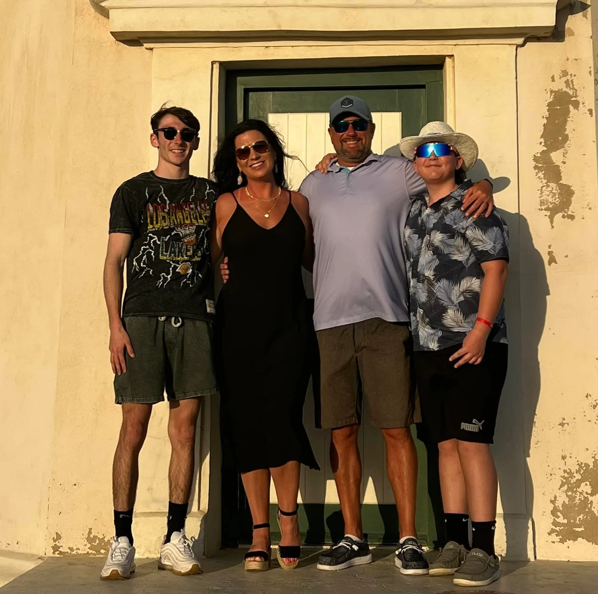 Four people standing close together outdoors in front of a green door, smiling, wearing casual summer clothing and sunglasses.