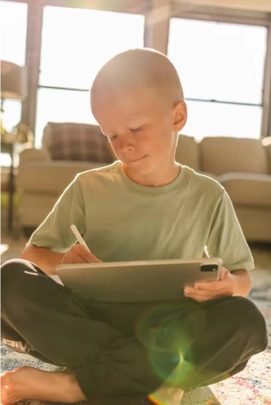 A young bald boy sitting cross-legged on the floor, writing or drawing on a tablet, with bright sunlight coming through windows behind him.