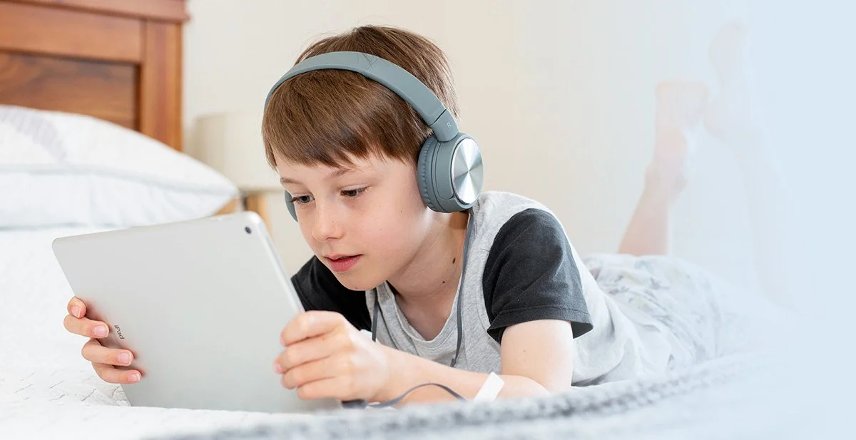 A young boy lying on his stomach on a bed, wearing headphones and looking at a tablet.