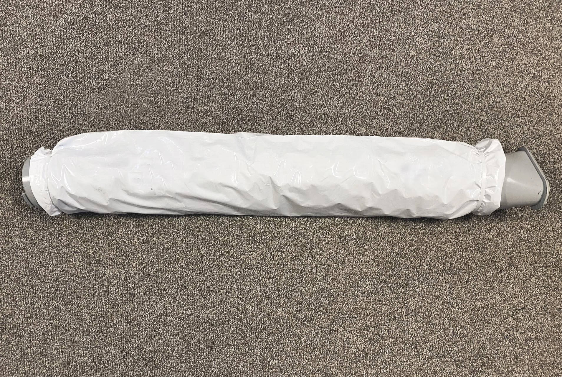 A cylindrical object wrapped in white plastic, lying on a brown, textured carpeted floor.
