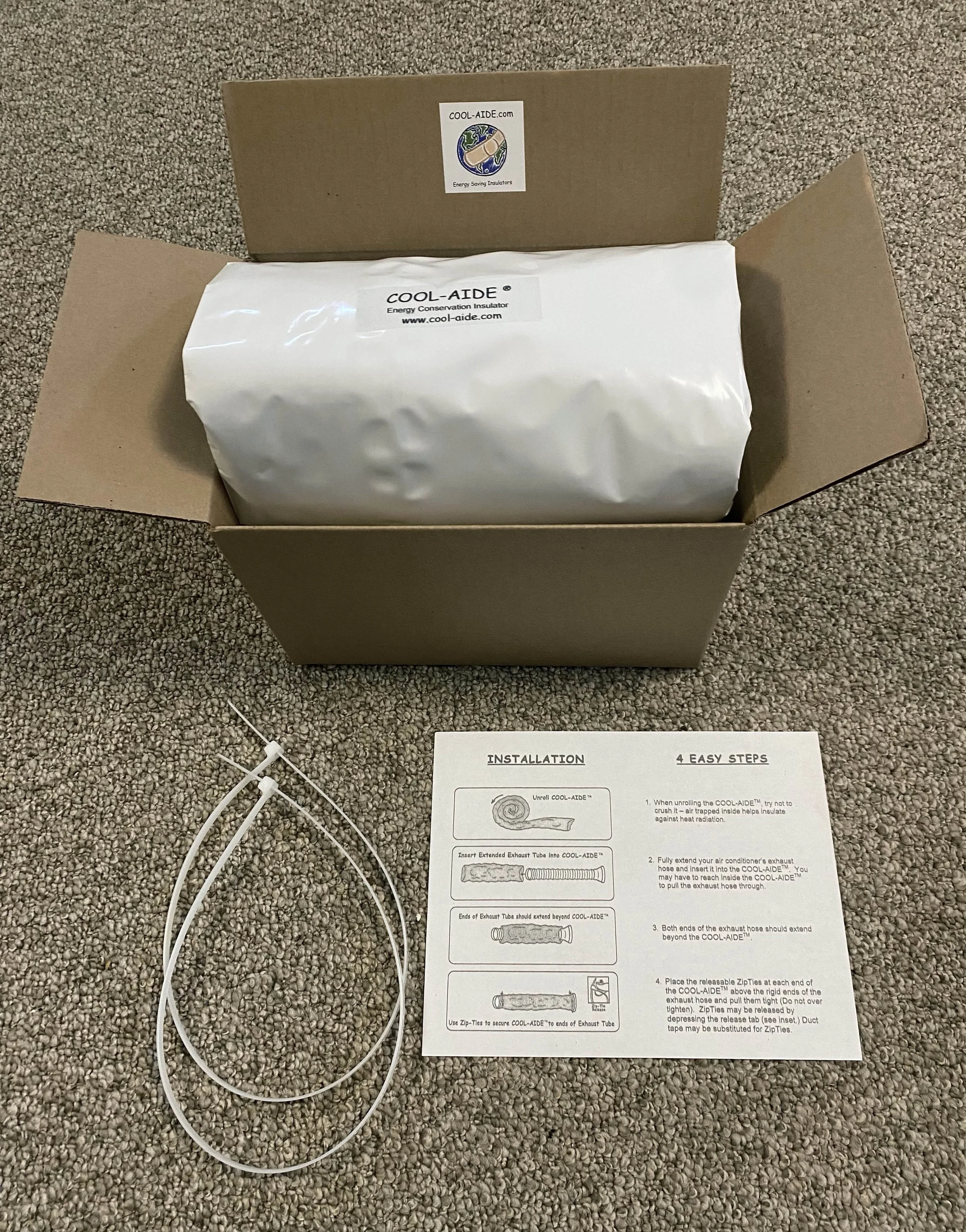 Open cardboard box containing a sealed white plastic bag with a label that reads "COOL-AIDE Energy Conservation Insulator". An instruction sheet with installation steps is laid out on a beige carpeted floor, along with two white zip ties and a partially visible white power cord.