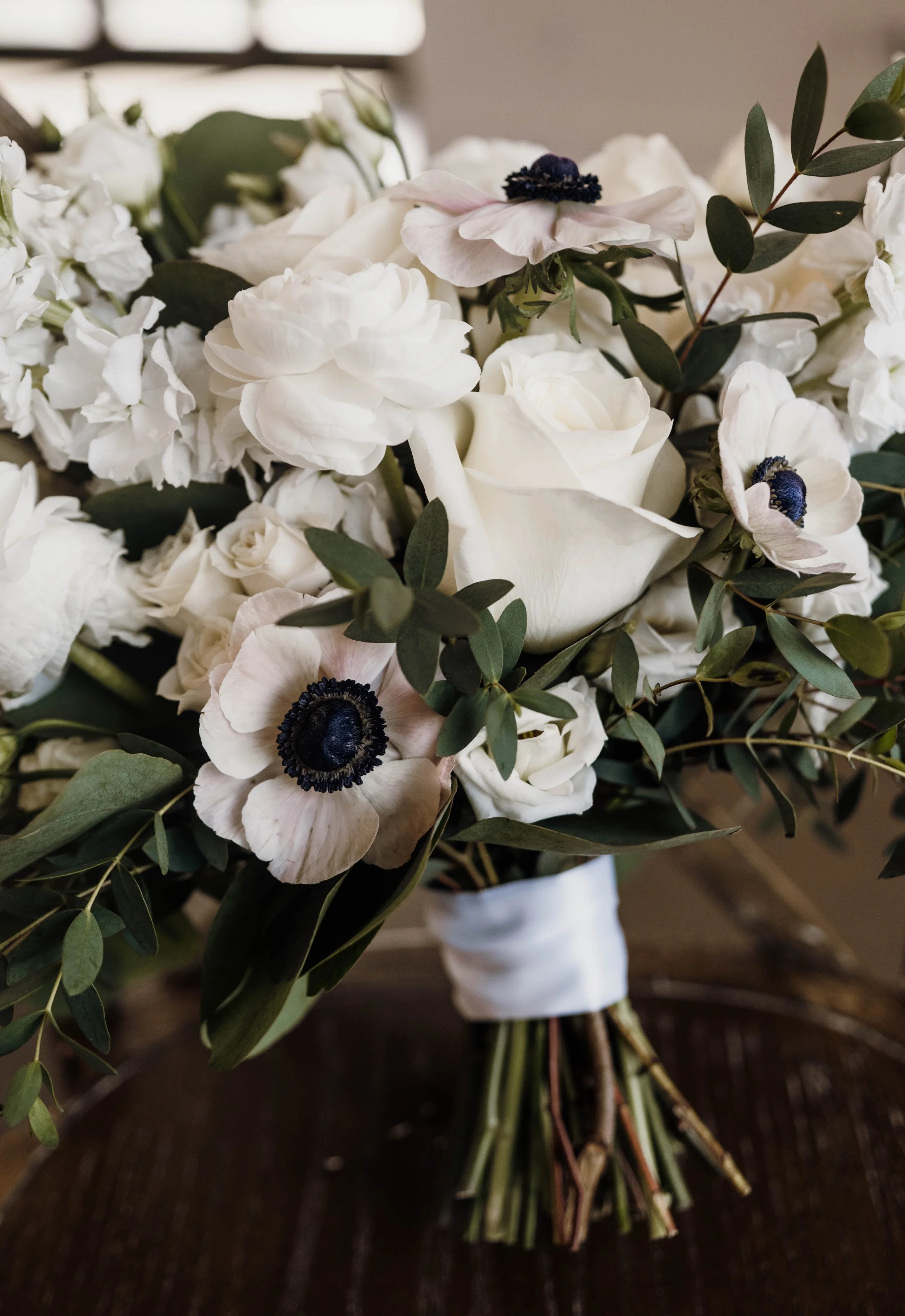 Close-up of a white floral bouquet with anemones, roses, and greenery, wrapped with white ribbon.