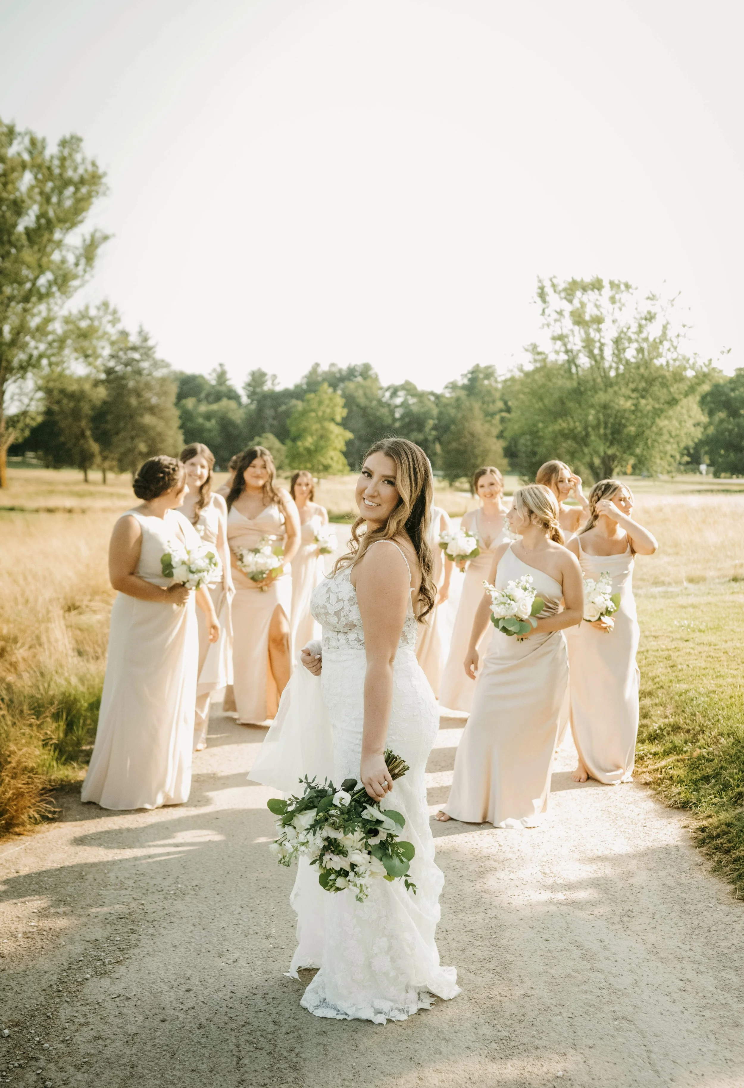 Bride in white wedding dress holding bouquet, surrounded by bridesmaids in pastel dresses with bouquets, outdoors on a sunny day with green trees in the background.