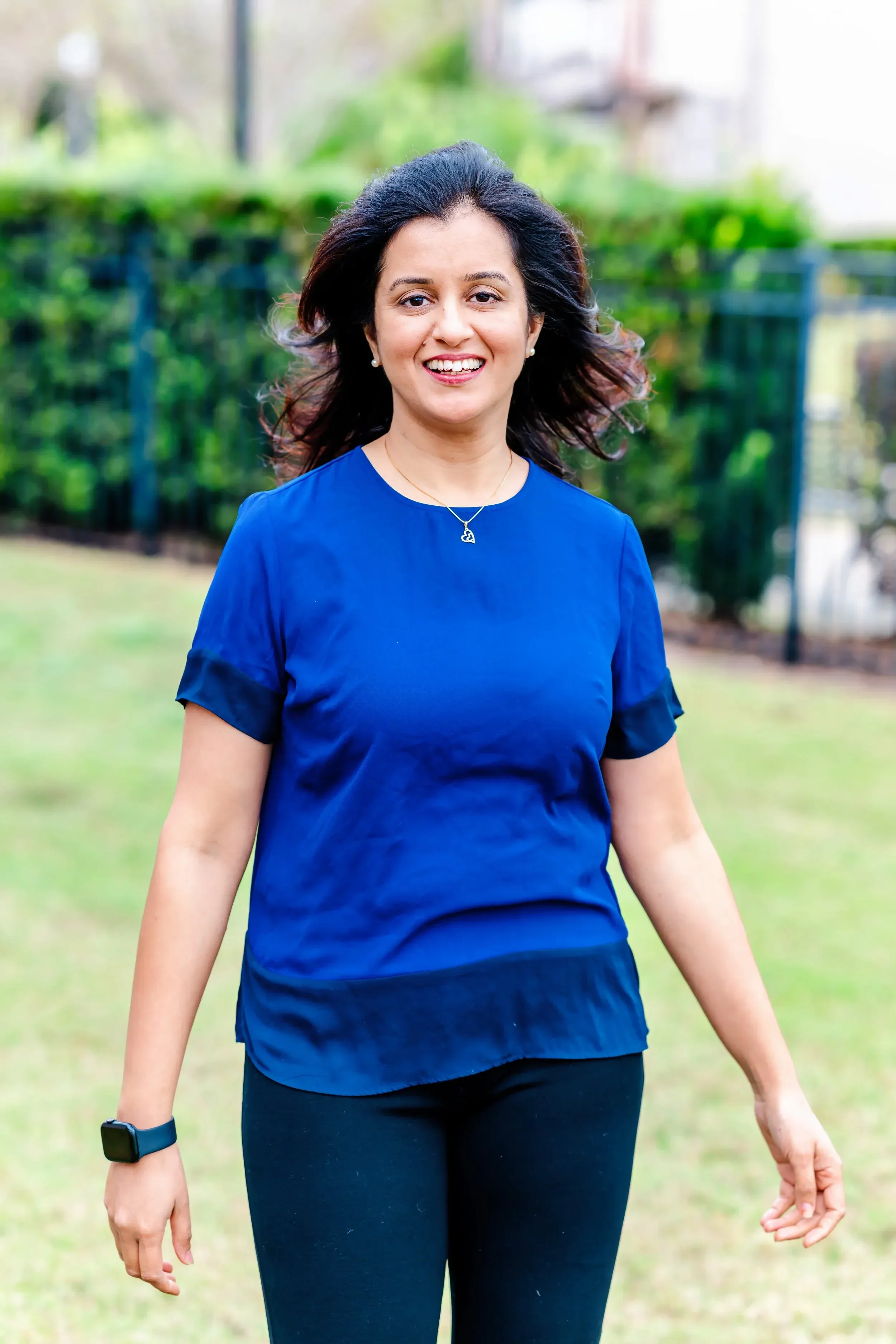 Dr. Rachita Bansal wearing a blue top and black leggings, smiling outdoors with green foliage in the background.