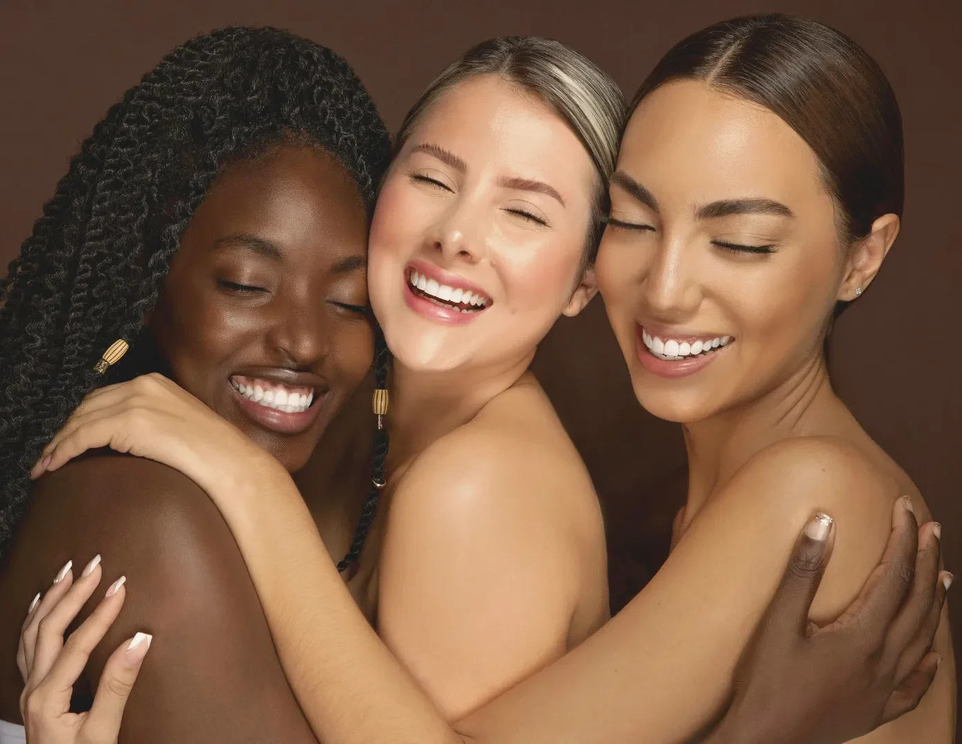 Three women of diverse ethnicities smiling and hugging each other against a brown background.