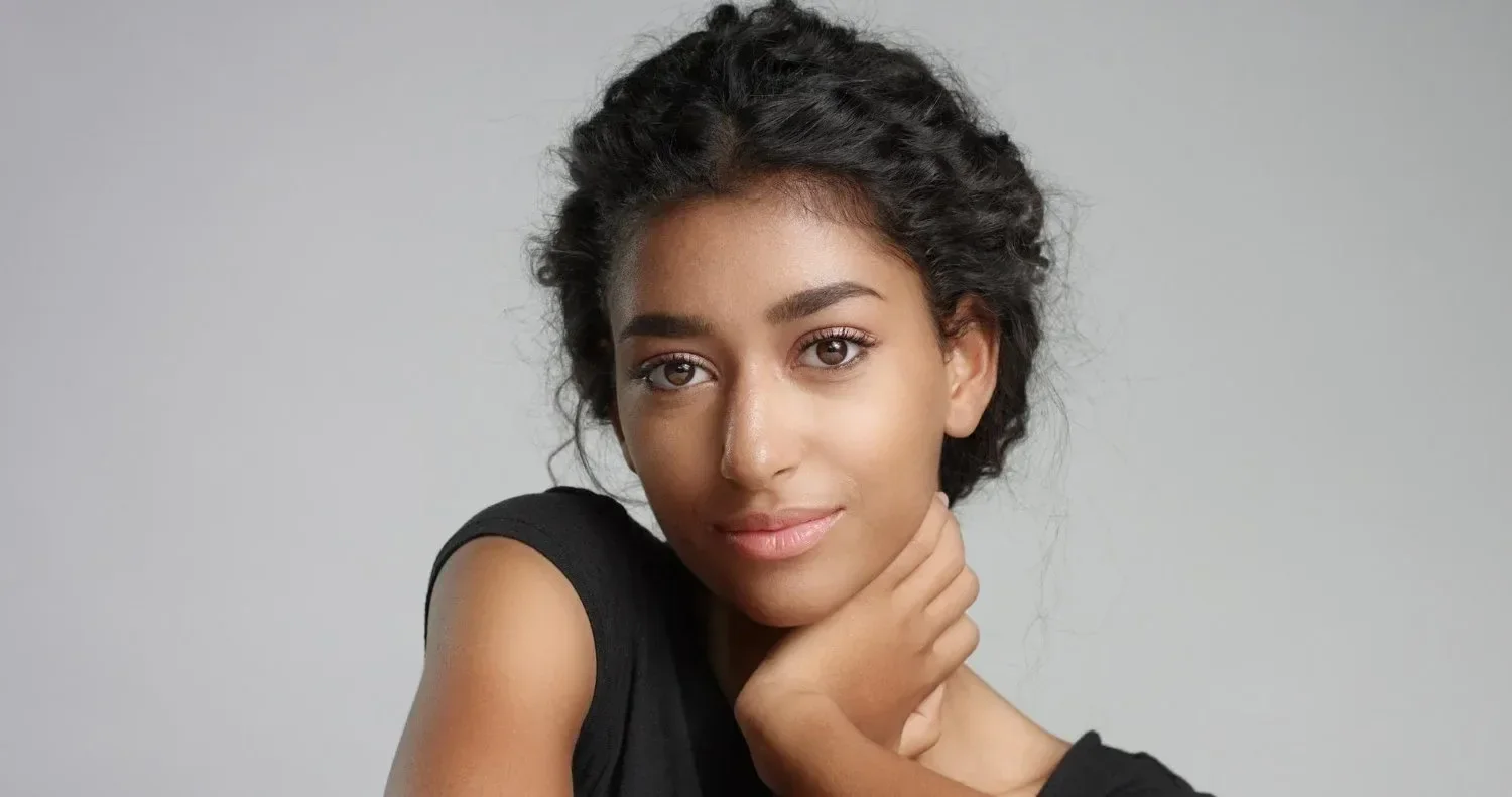 A young woman with dark wavy hair and light brown skin, wearing a black sleeveless top, gently holding her chin with her left hand, looking into the camera with a soft smile against a plain gray background.