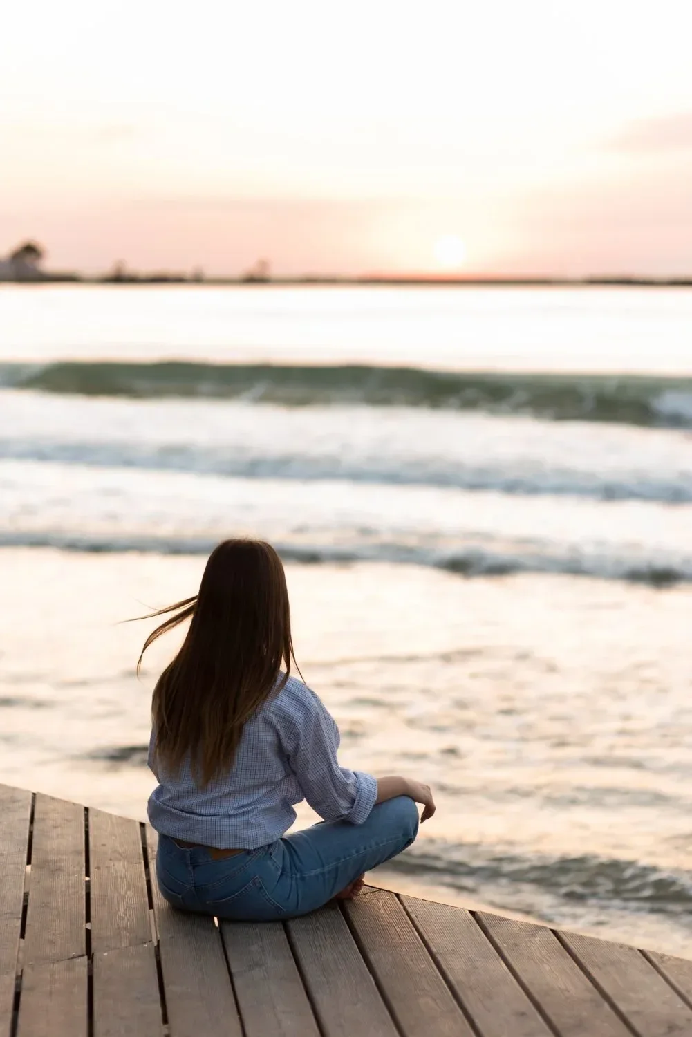 A woman with long brown hair, wearing a blue shirt and jeans, sitting barefoot on a wooden deck by the water at sunset, gazing at the ocean.