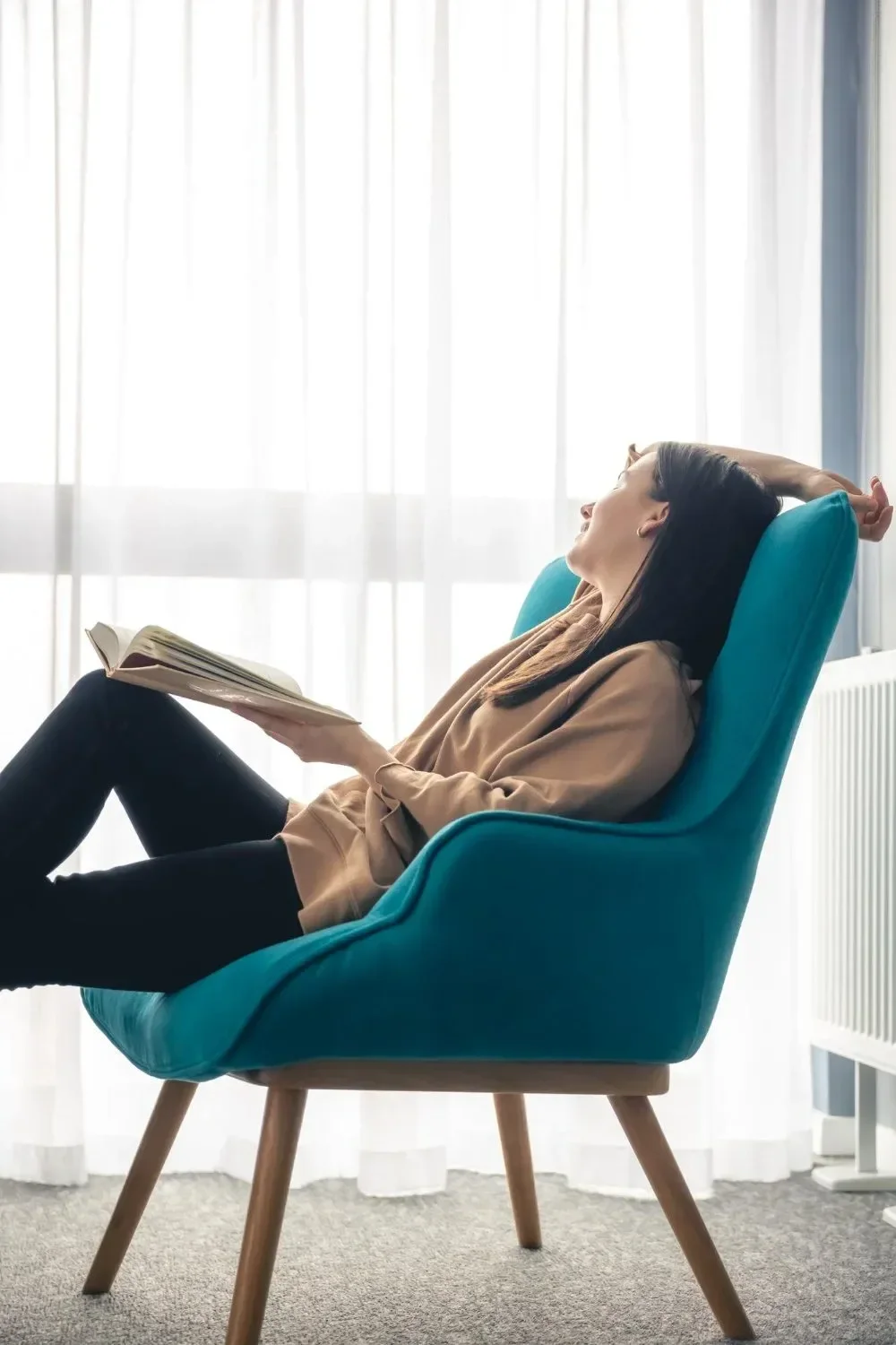 A woman sitting in a teal armchair reading a book, with her arm resting behind her head, in a bright room with white curtains.