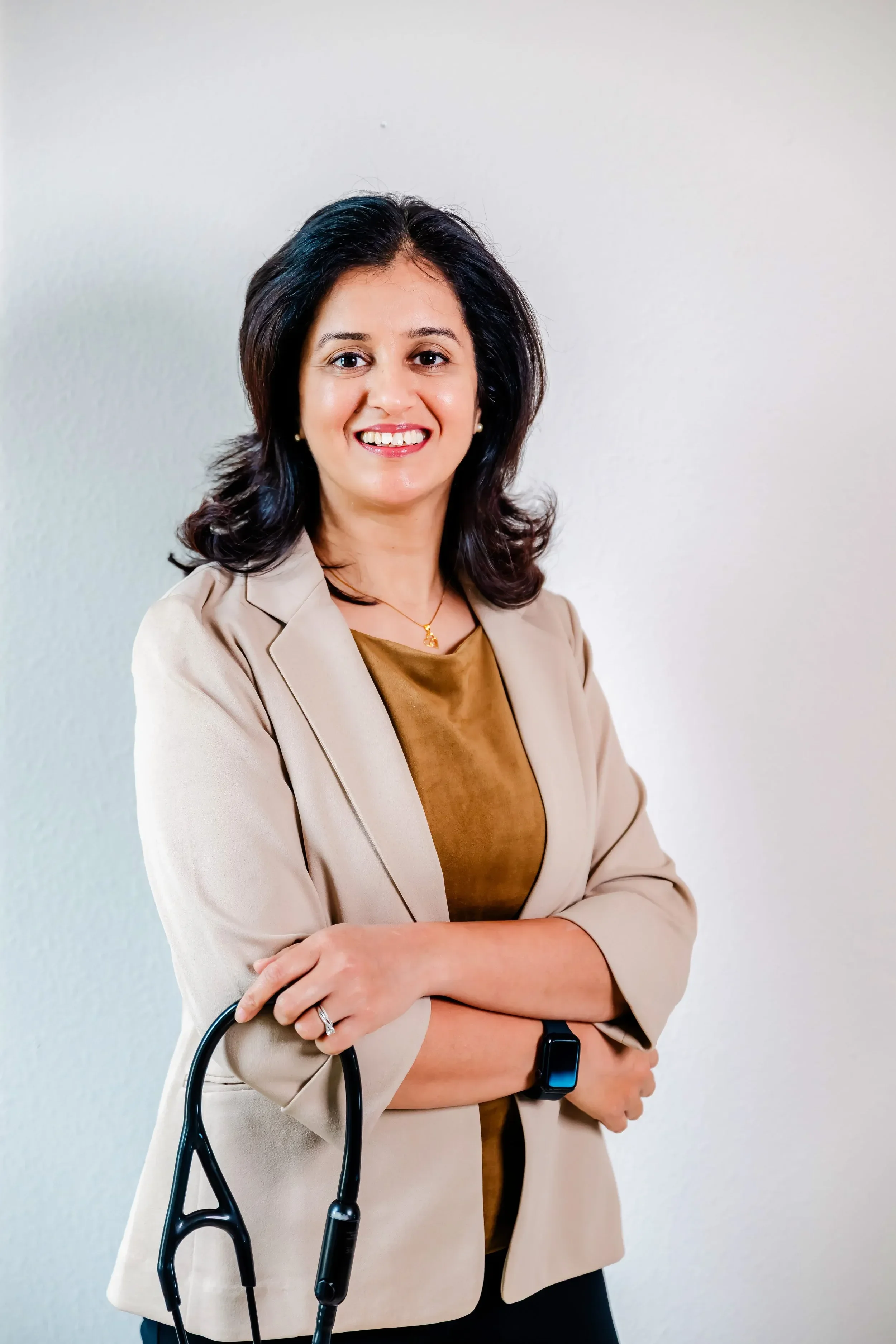 Dr. Rachita Bansal with black hair, wearing a beige blazer and a brown top, holding a stethoscope and wearing a smartwatch, standing against a light-colored background.