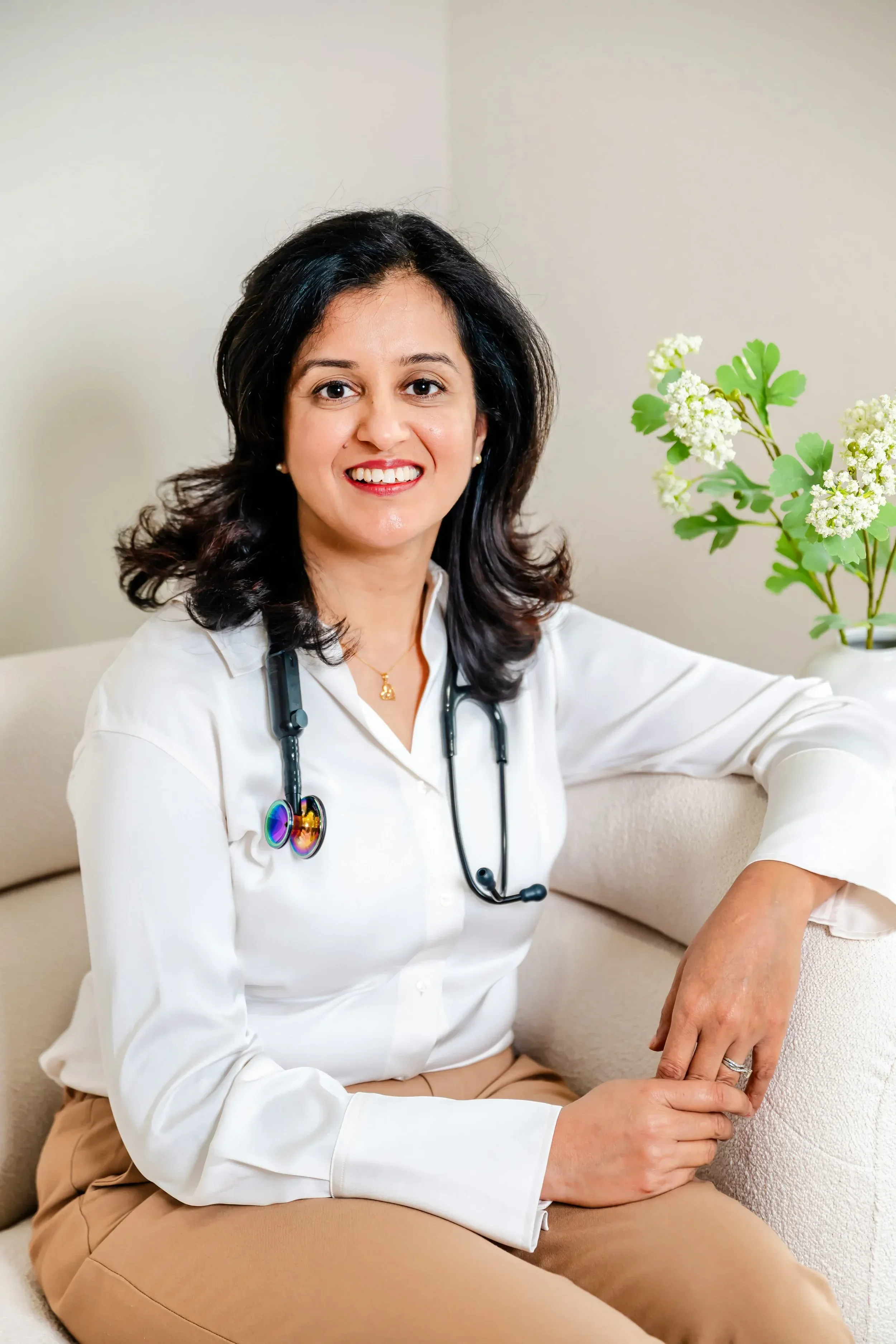 Dr. Rachita Bansal, wearing a white coat and stethoscope, sitting on a sofa with a colleague's hand on her knee, in a room with a beige wall and a plant with white flowers nearby.