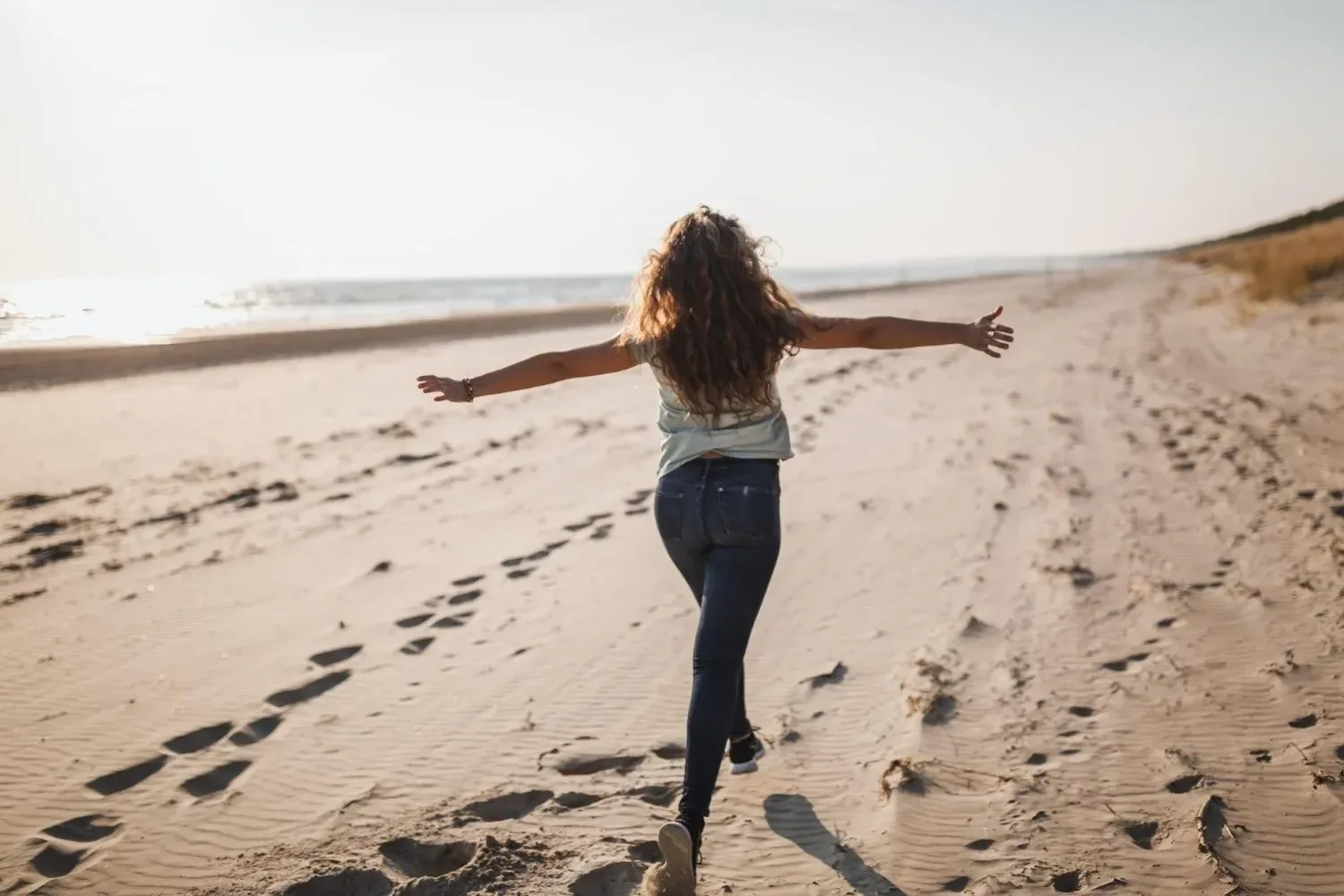 A woman with long curly hair running on a sandy beach with her arms outstretched and footprints in the sand, near the ocean during sunset.