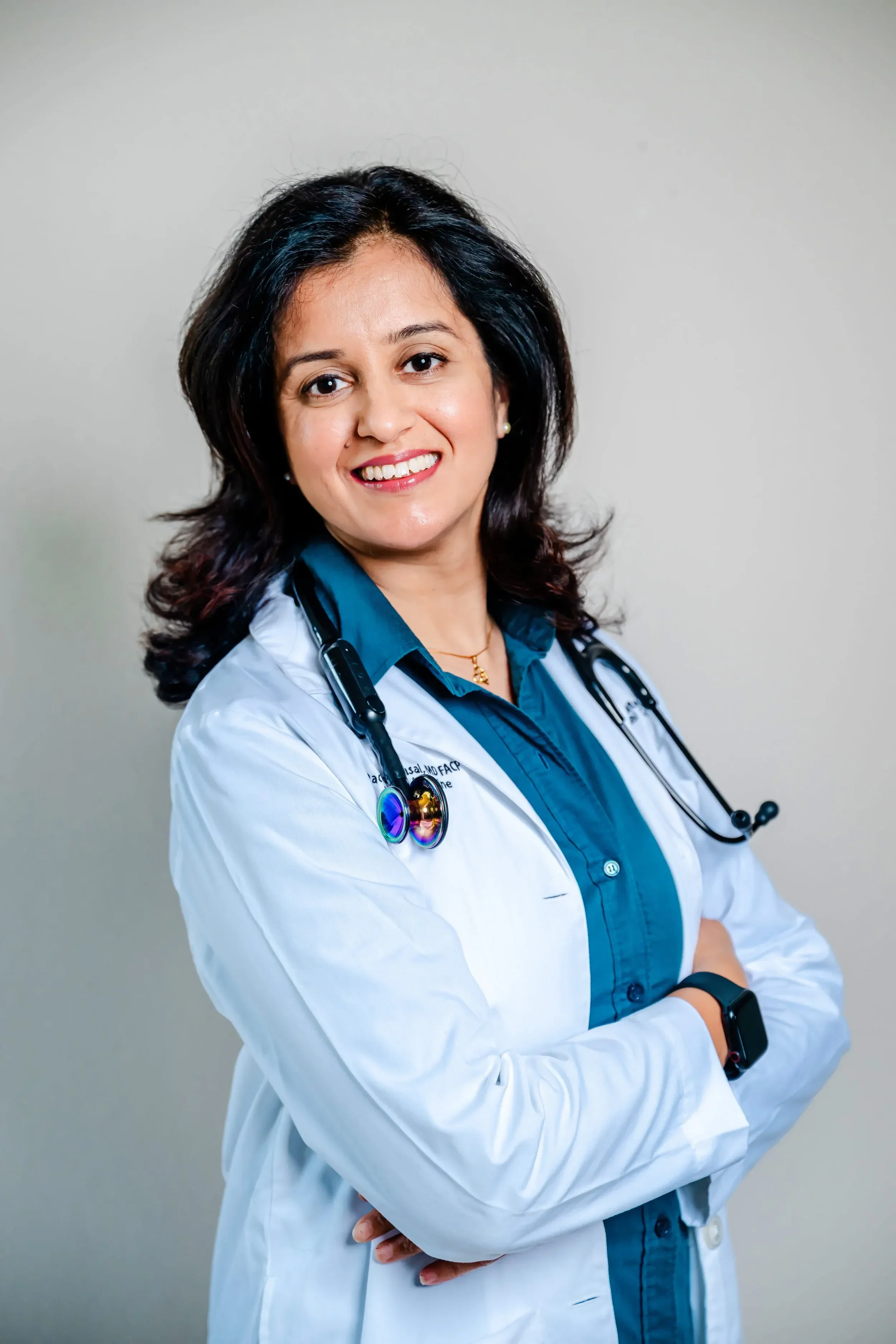 Dr. Rachita Bansal with black hair, wearing a white lab coat and stethoscope, standing with arms crossed against a plain background.